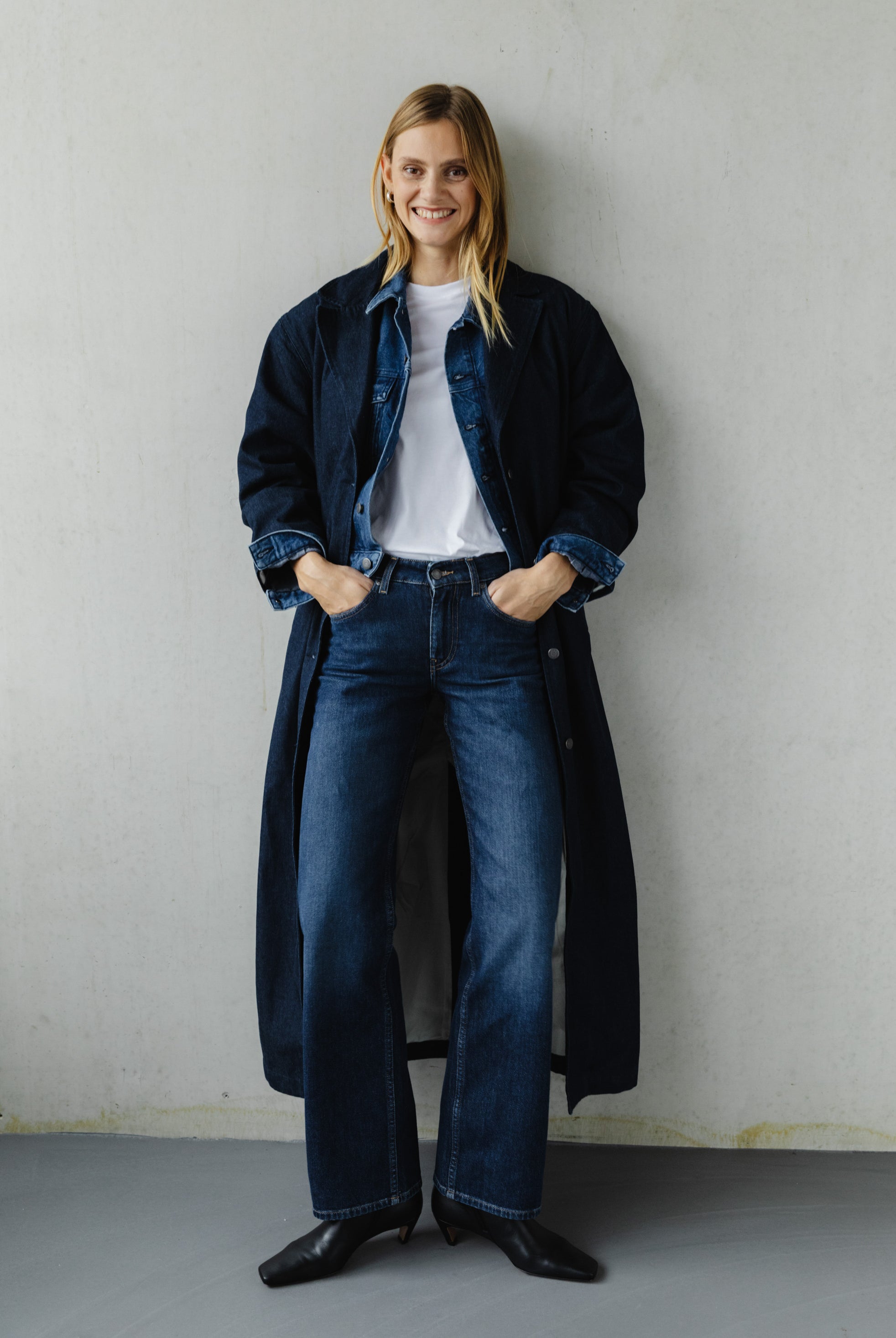 Woman wearing dark blue denim jeans and denim long coat, paired with a white tee, standing against a plain wall.