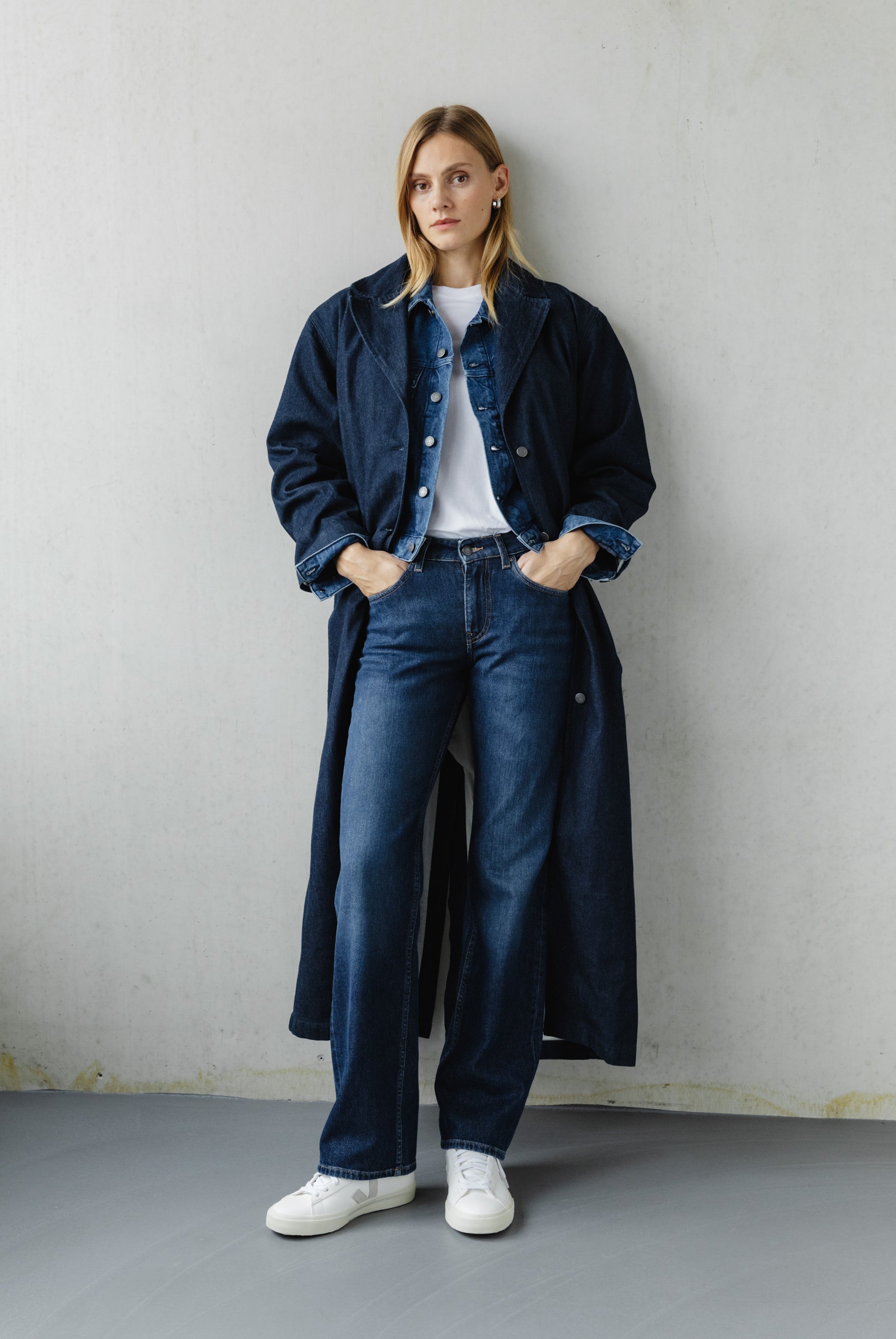 Woman wearing a stylish denim ensemble with a long coat, paired with a white t-shirt and sneakers, leaning against a wall.