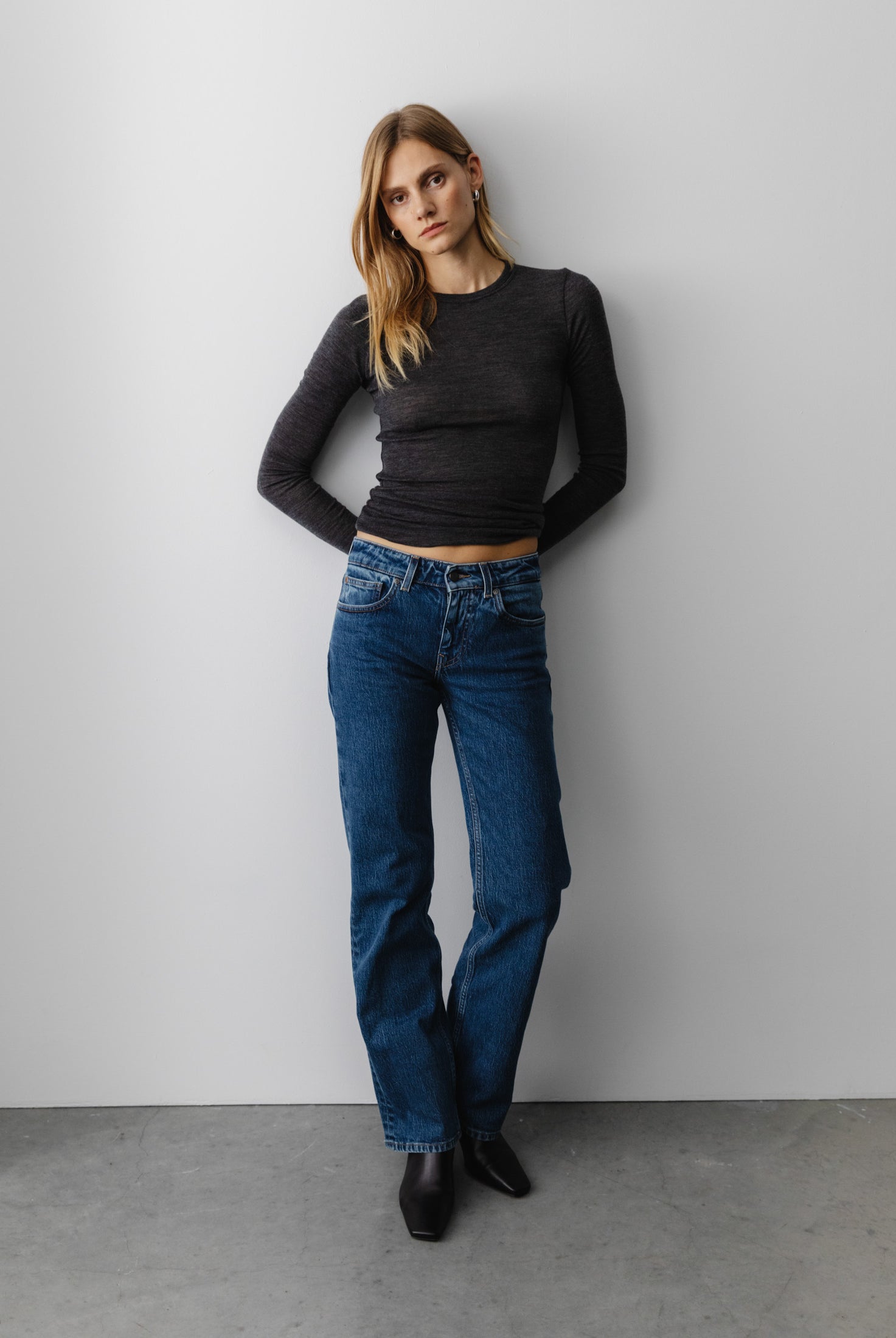 Model in dark gray long-sleeve top and blue straight-leg jeans, standing against a minimalist white wall.
