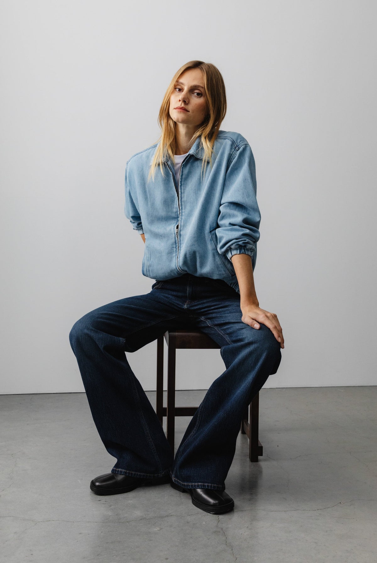 Casual fashion portrait with light blue denim jacket and dark blue flared jeans, seated on a wooden stool against a plain backdrop.