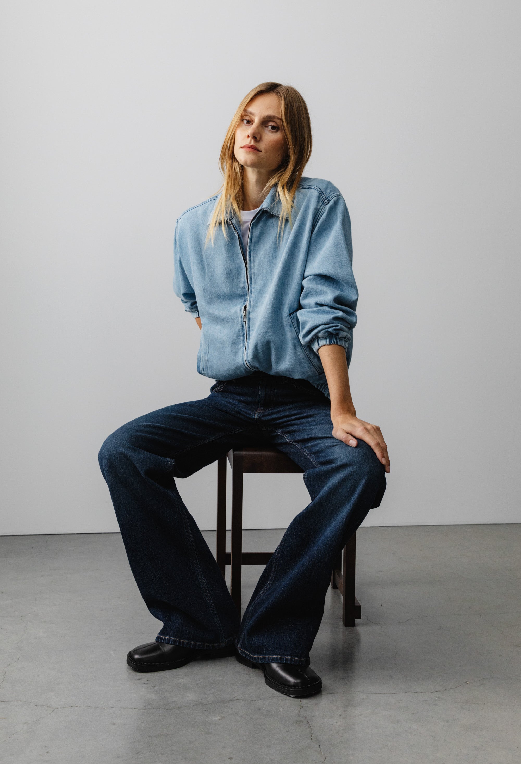 Casual fashion portrait with light blue denim jacket and dark blue flared jeans, seated on a wooden stool against a plain backdrop.