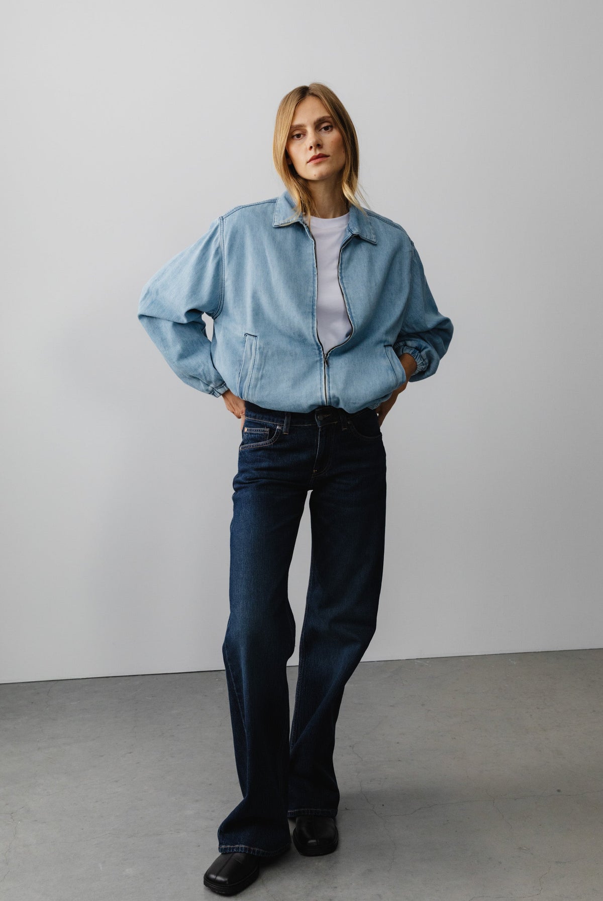 Model wearing a light blue denim jacket with a white shirt and dark blue jeans, standing in a minimalistic setting.