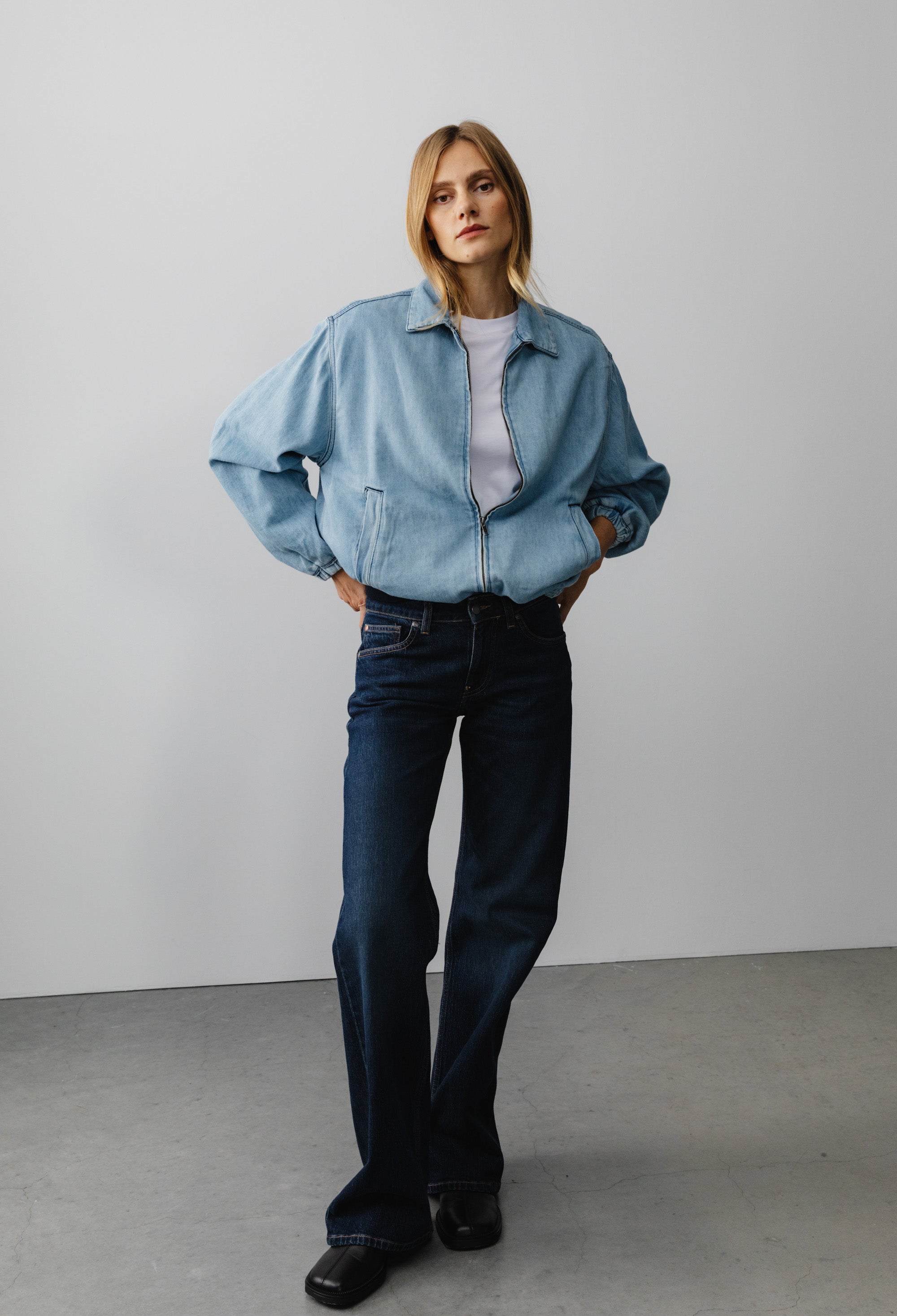 Model wearing a light blue denim jacket with a white shirt and dark blue jeans, standing in a minimalistic setting.
