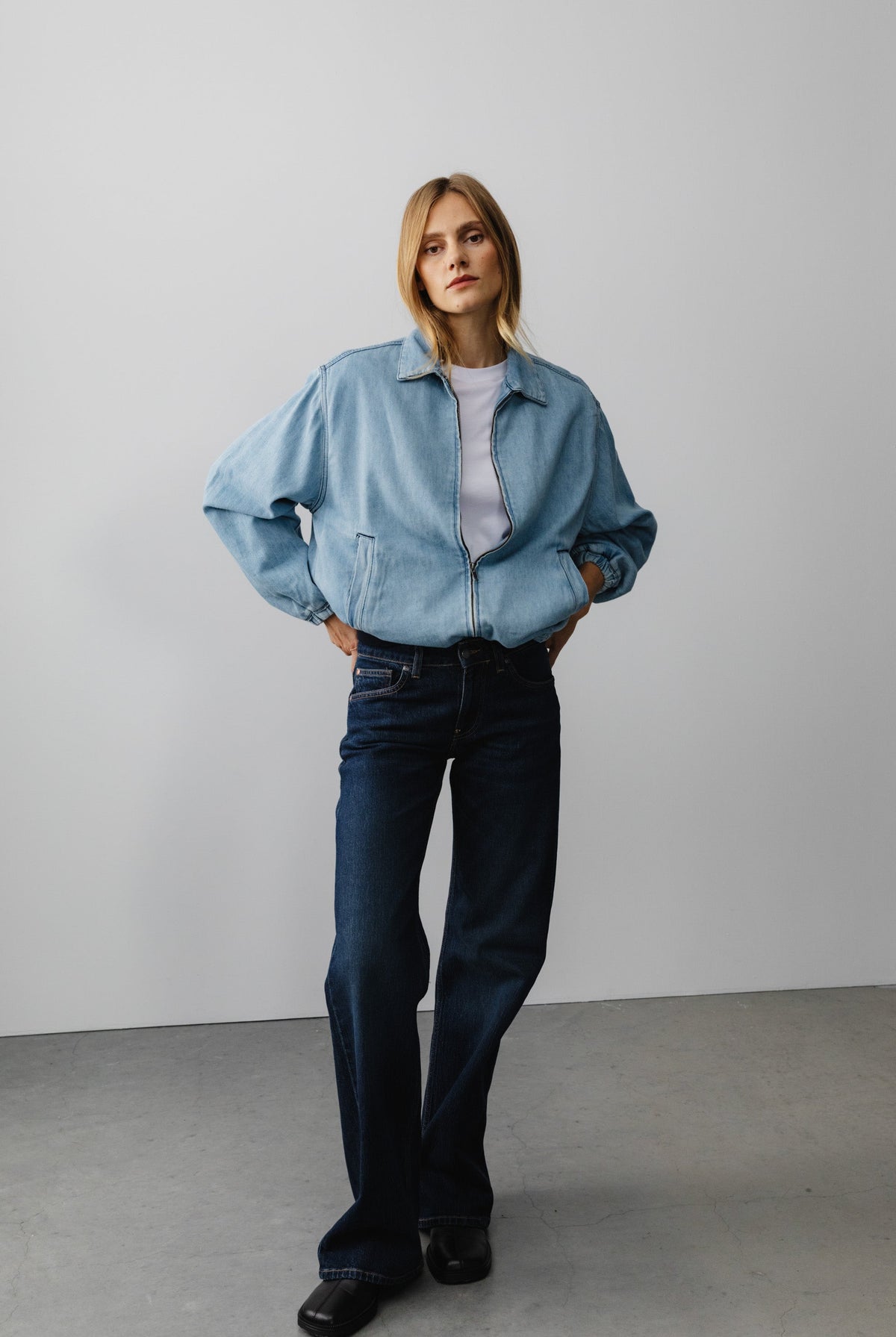 Woman in light blue, oversized denim jacket with dark blue, wide-leg jeans and black shoes, standing against gray wall.