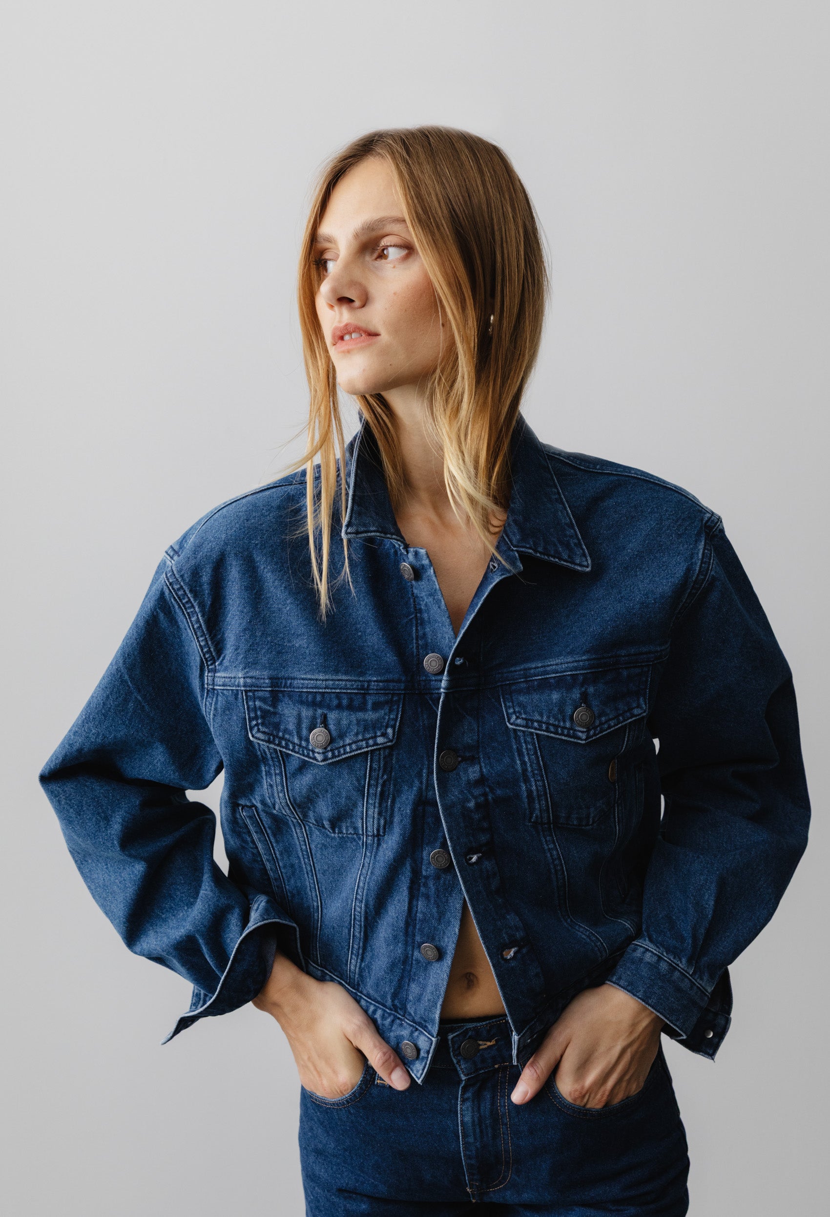 Casual denim jacket in deep indigo worn by a person with hands in pockets, featuring button details and a classic collar.