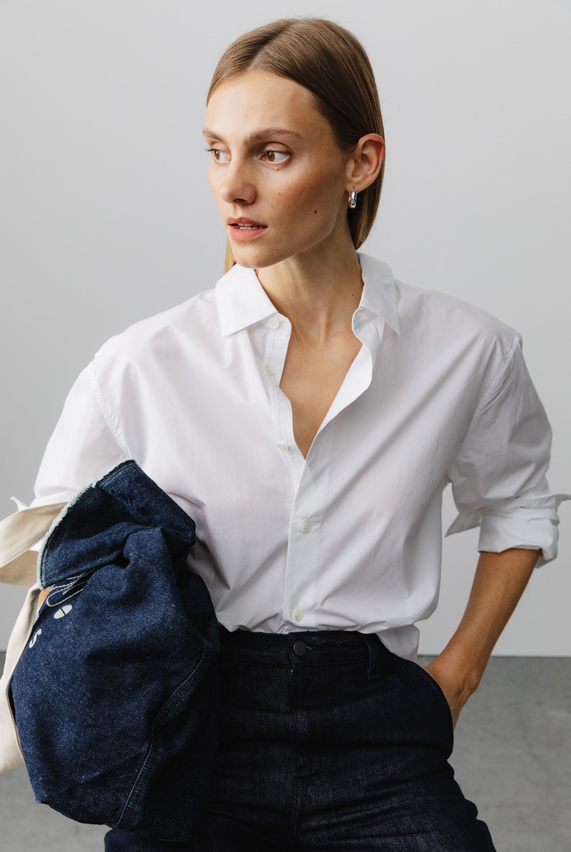 Woman wearing a crisp white button-up shirt with rolled sleeves, paired with dark indigo denim jeans, holding a navy jacket.