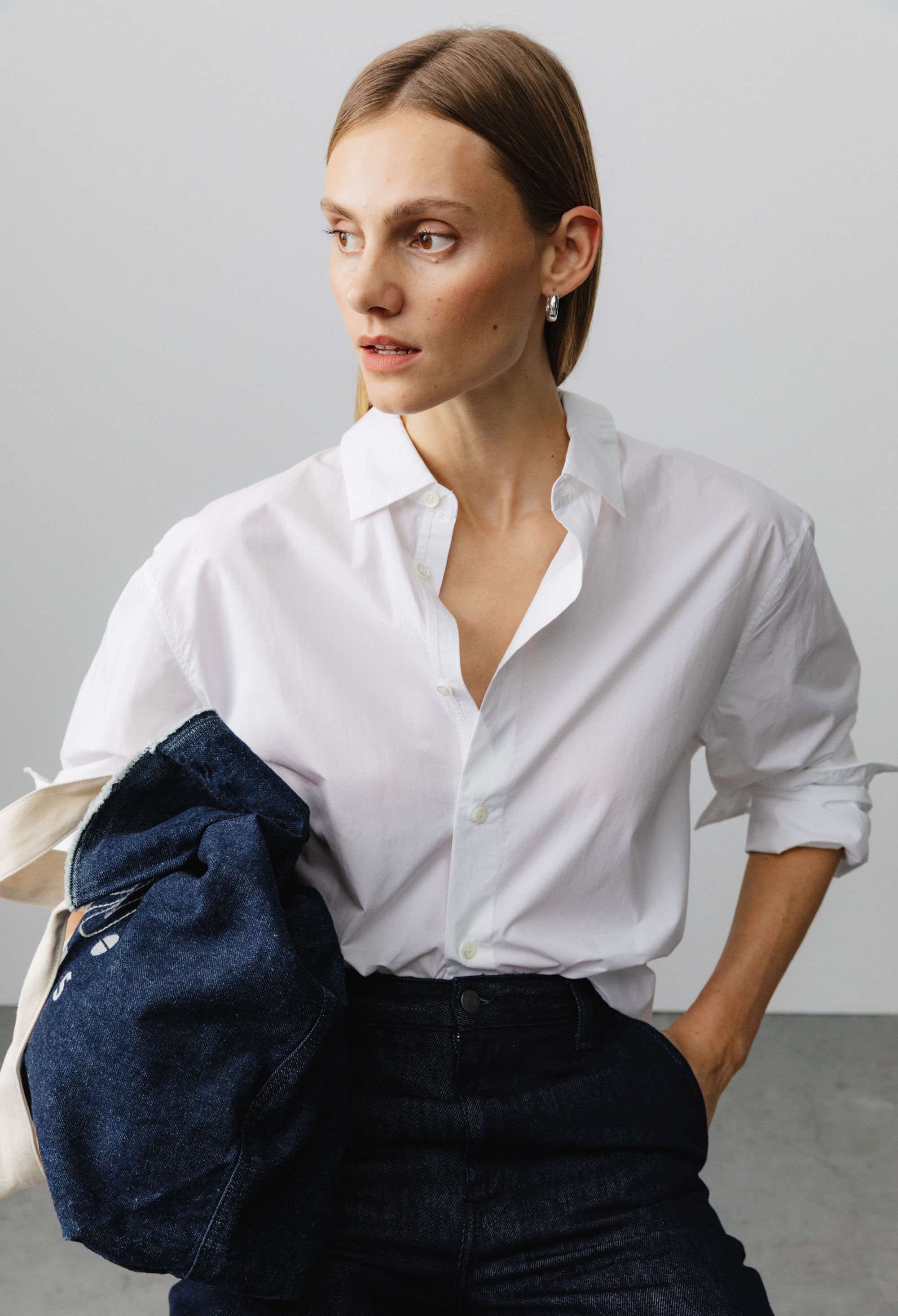 Woman wearing a crisp white button-up shirt with rolled sleeves, paired with dark indigo denim jeans, holding a navy jacket.