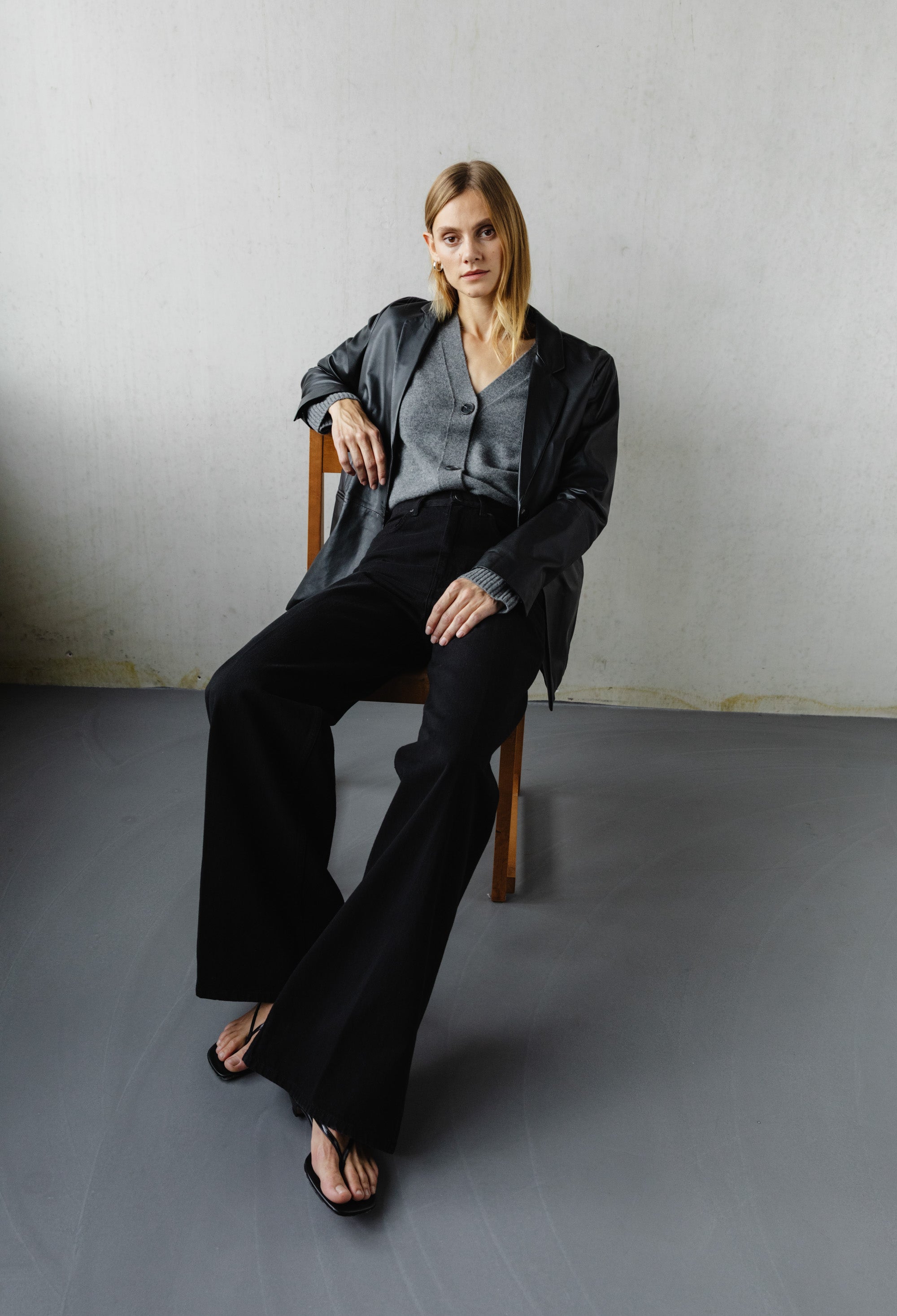 Stylish woman in a gray cardigan and black wide-leg pants, seated on a wooden chair against a minimalist concrete wall.