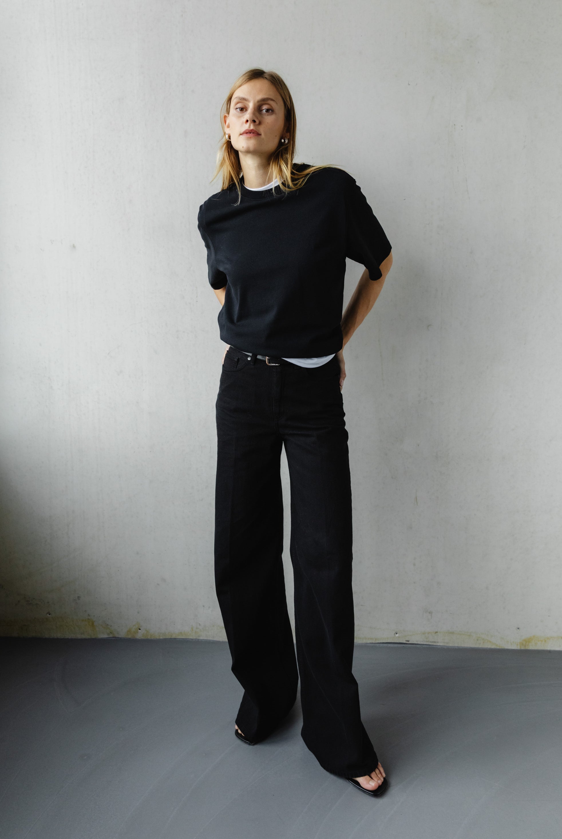 A woman models a minimalist outfit of a black t-shirt and wide-leg black trousers in a neutral indoor setting.