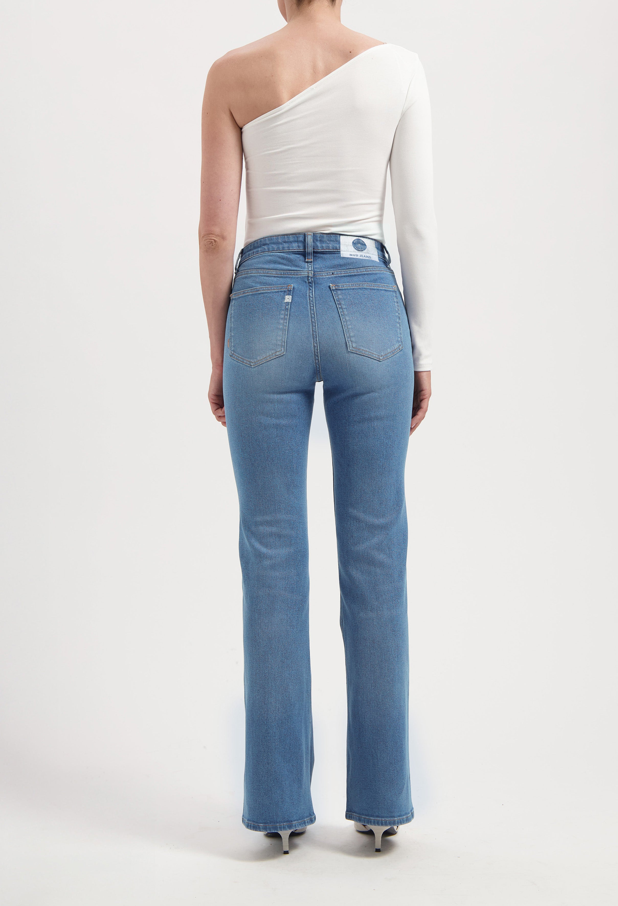 Back view of light blue, high-waisted MUD Jeans in organic cotton, worn with a white, one-shoulder long-sleeve top.