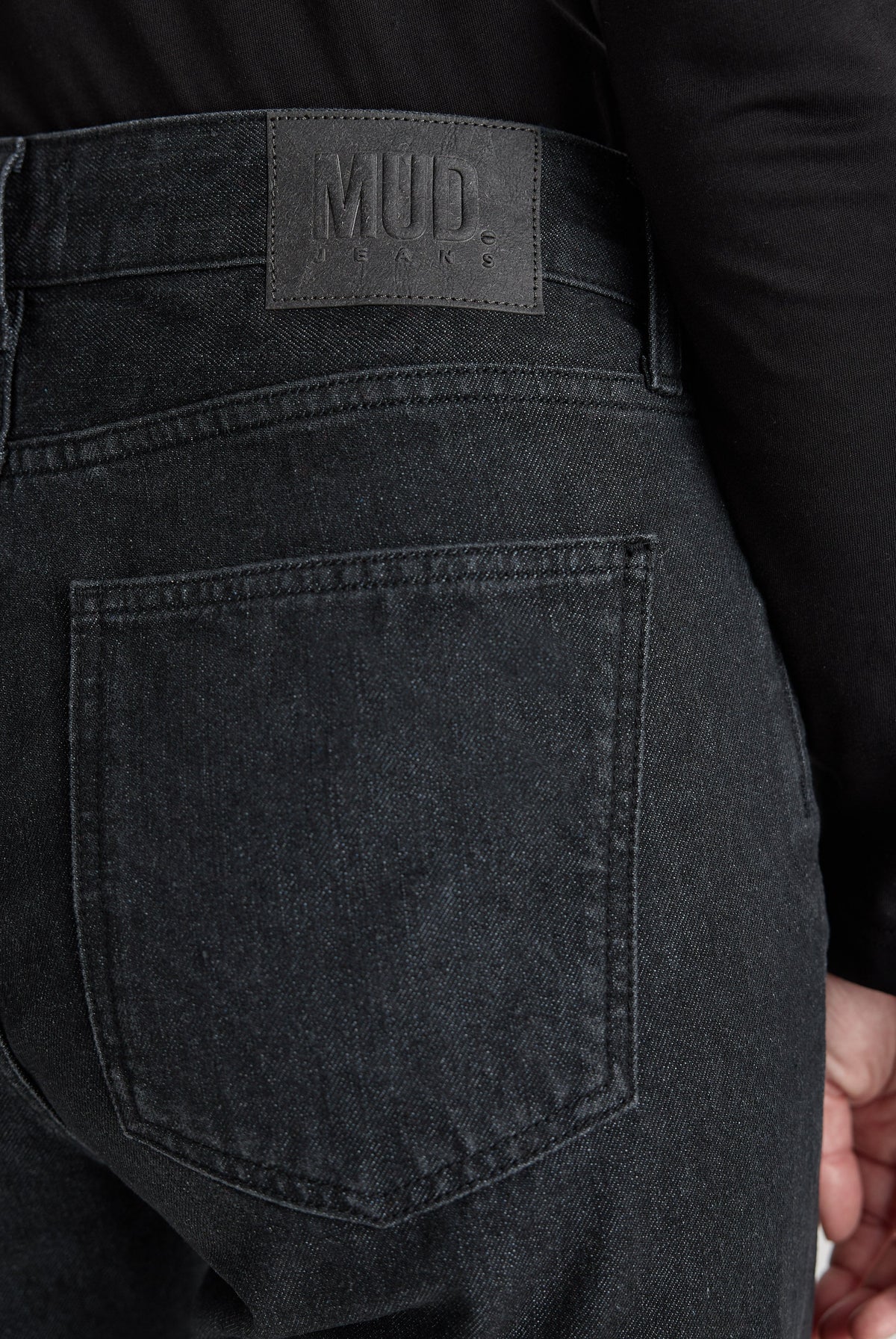 Close-up of MUD Jeans featuring sustainable dark gray denim, showcasing back pocket and leather label, worn with black top.