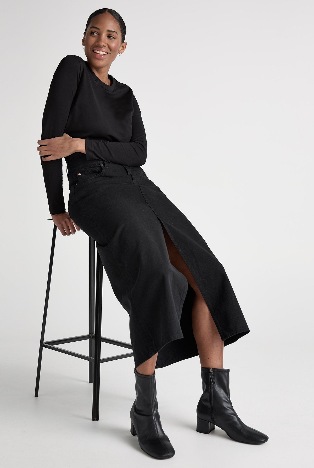 Woman in a stylish black long-sleeve shirt, black denim skirt, and ankle boots seated on a minimalist stool against a white background.