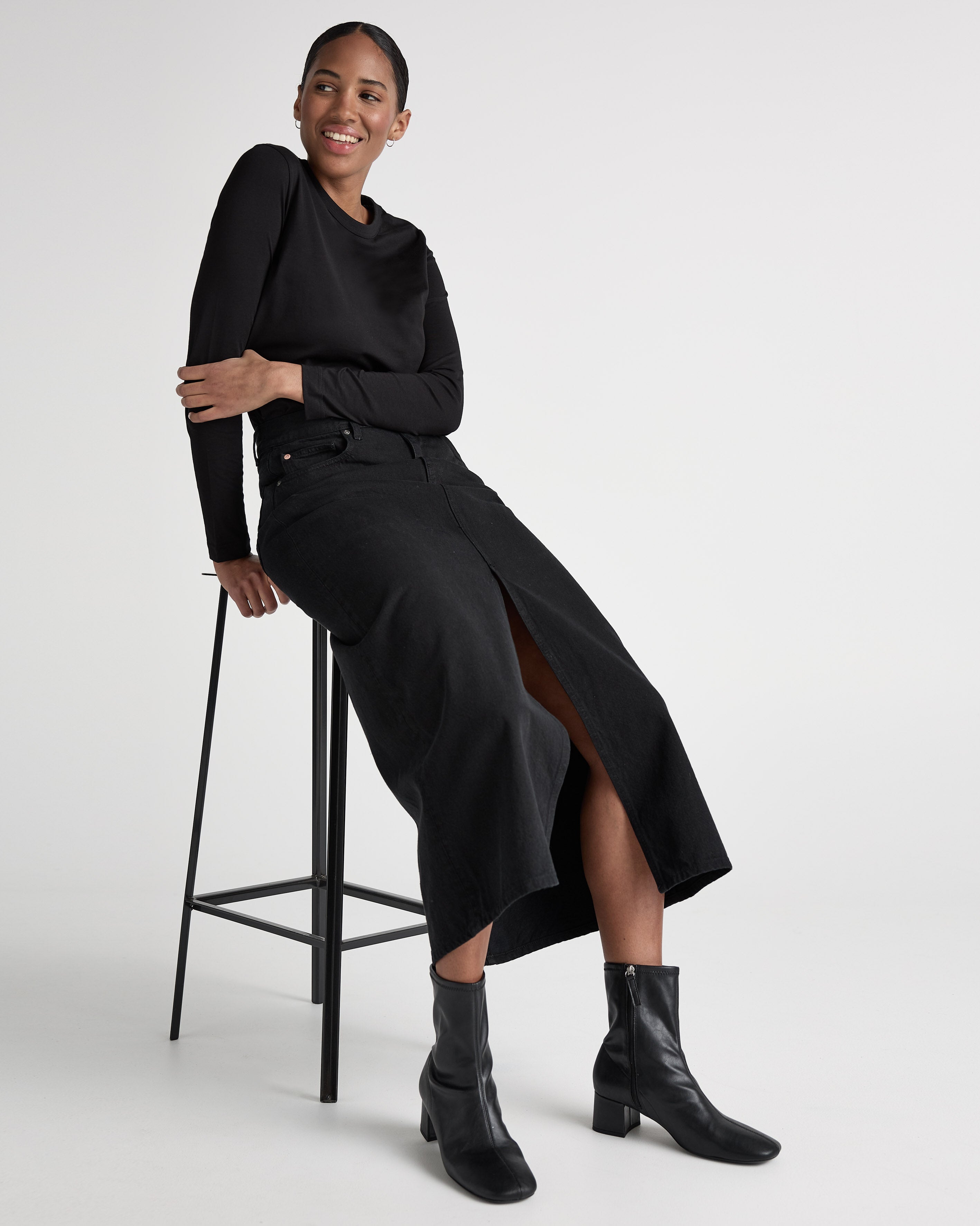 Woman in a stylish black long-sleeve shirt, black denim skirt, and ankle boots seated on a minimalist stool against a white background.