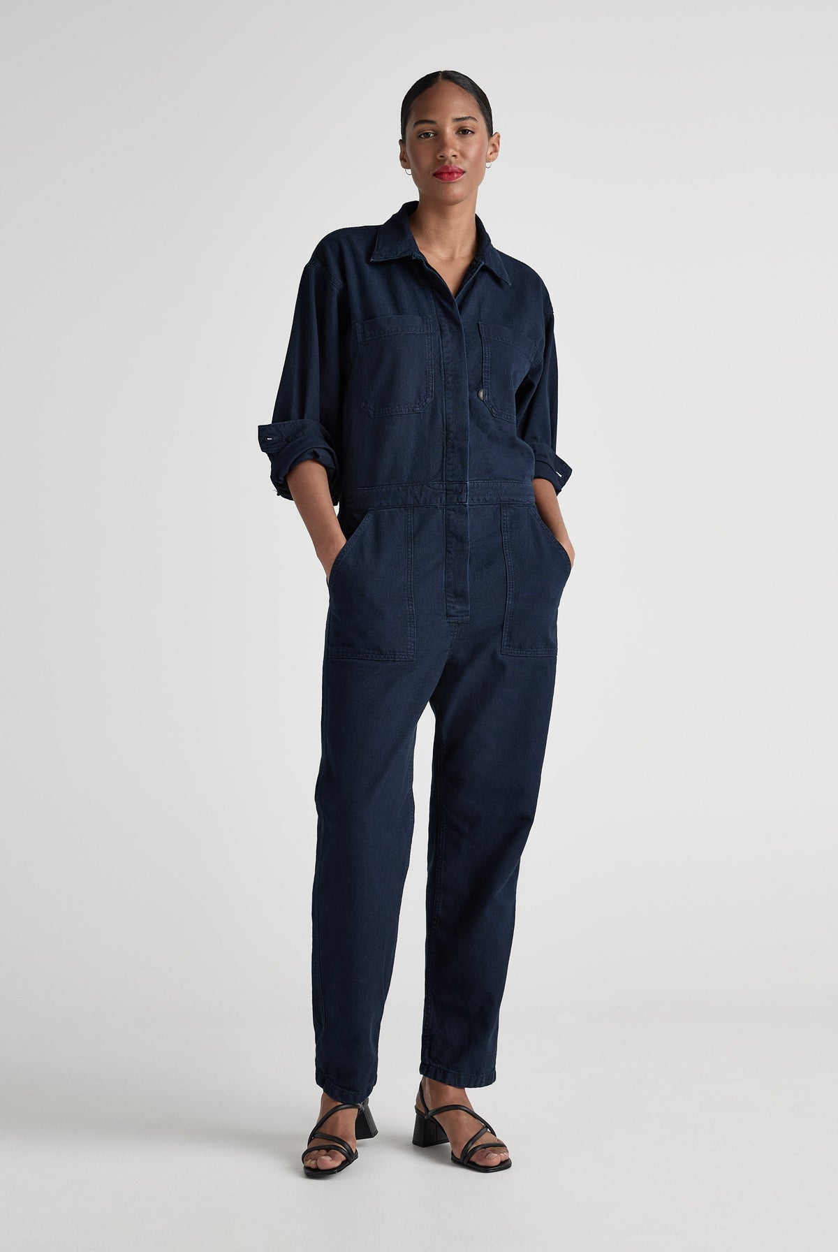 Navy blue denim jumpsuit with long sleeves and button-front design, modeled with confidence and paired with black sandals.