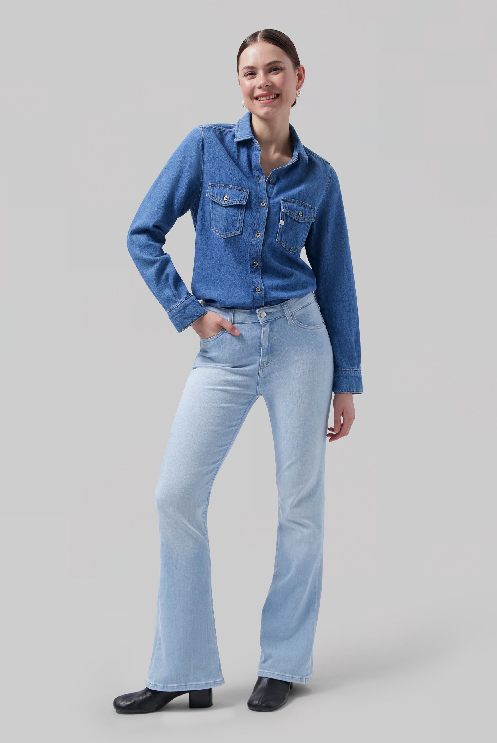 A woman wearing a classic blue denim shirt with front pockets paired with light blue flared jeans and black ankle boots.