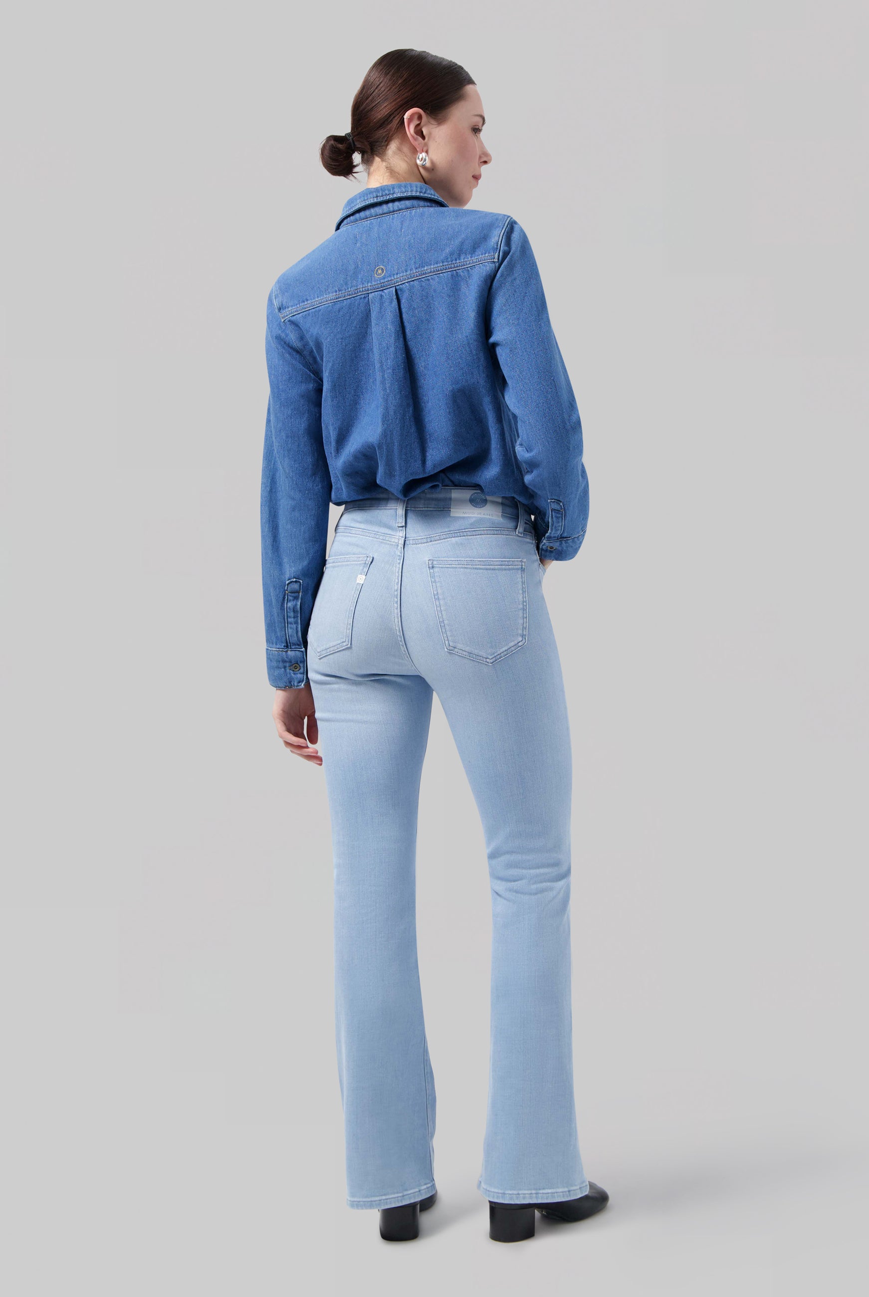 Woman wearing light blue flared jeans and a long-sleeve denim shirt, viewed from the back, styled with earrings.