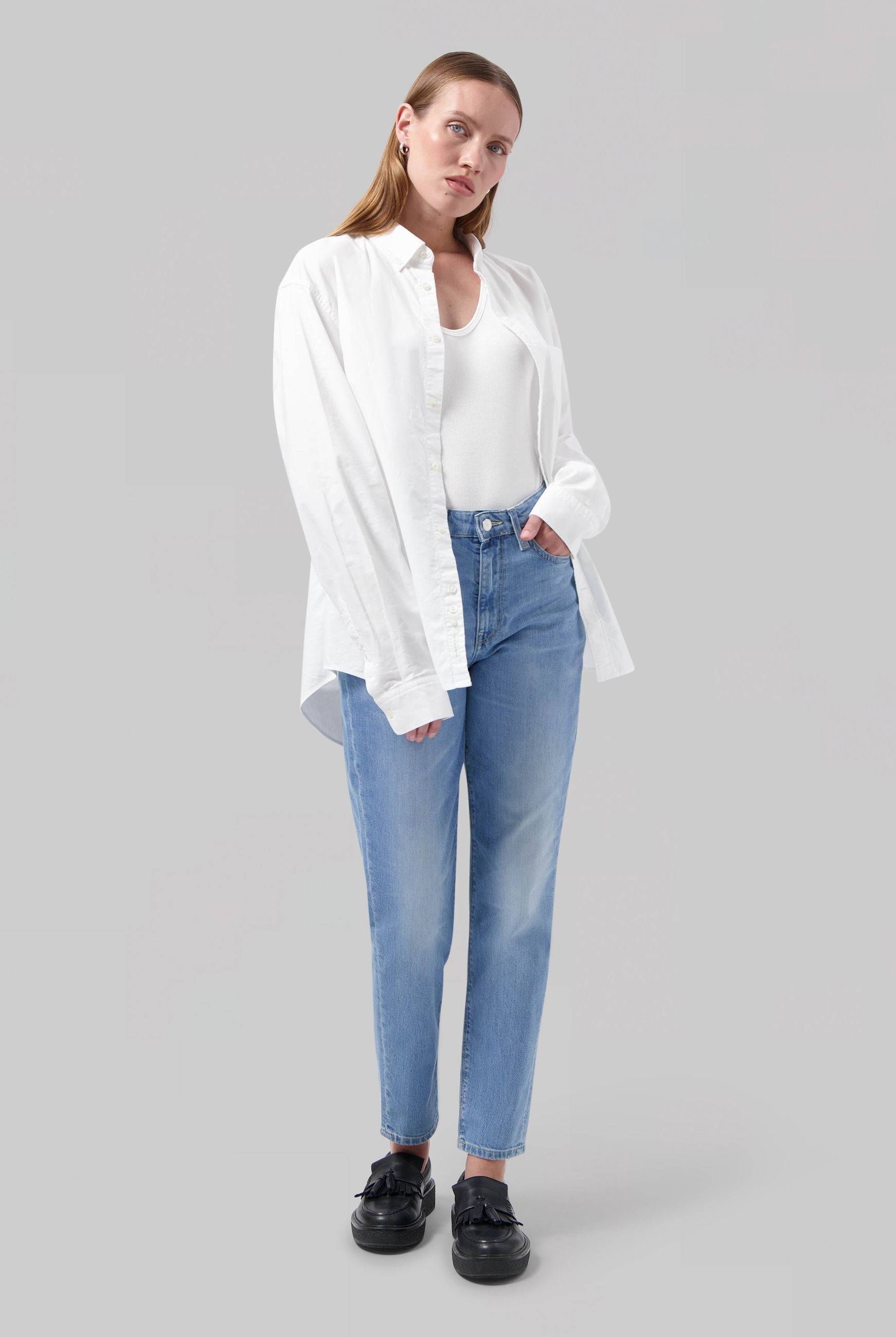 Casual women’s outfit featuring a white shirt over a white top and light blue high-waisted jeans, paired with black loafers.
