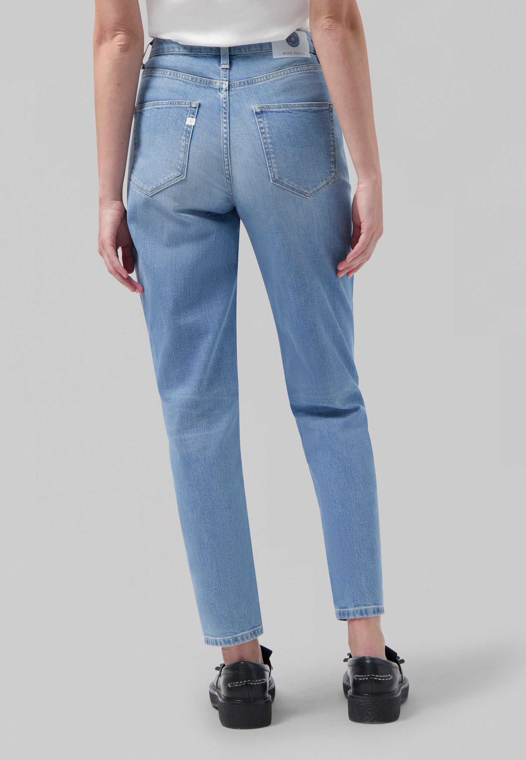 Light blue sustainable denim jeans from MUD Jeans, featuring high-waisted, straight-leg fit worn with black loafers and a white top.