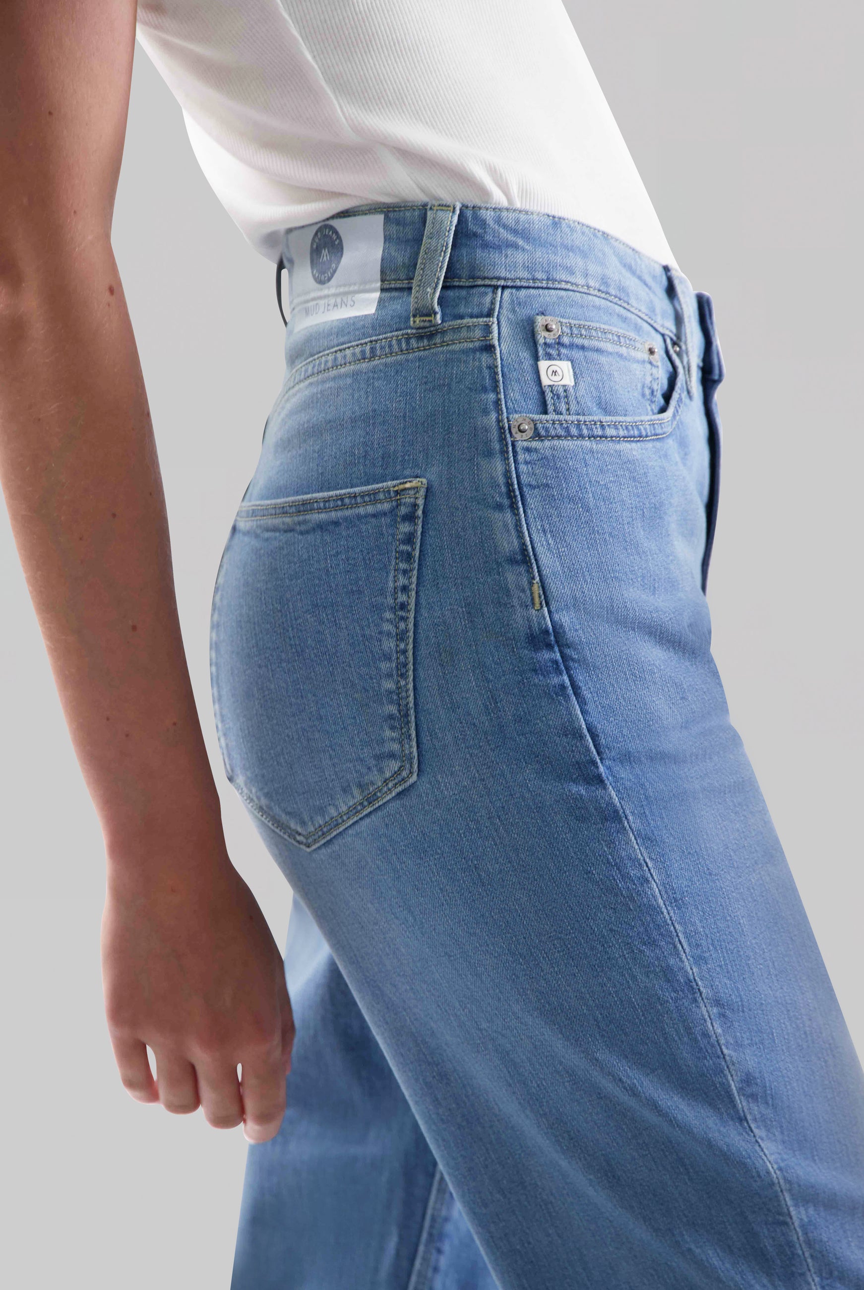 High-waisted light blue denim jeans from MUD Jeans, featuring a side view fit and paired with a white sleeveless top.