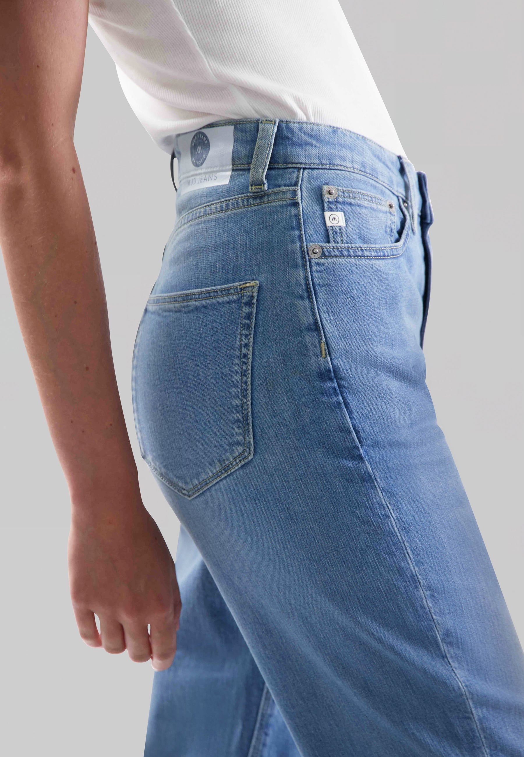 High-waisted light blue denim jeans from MUD Jeans, featuring a side view fit and paired with a white sleeveless top.