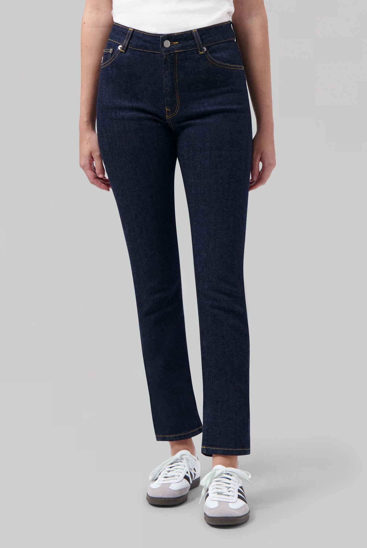 Dark blue high-waisted denim jeans with contrast stitching, worn with a white ribbed tank top and white sneakers.