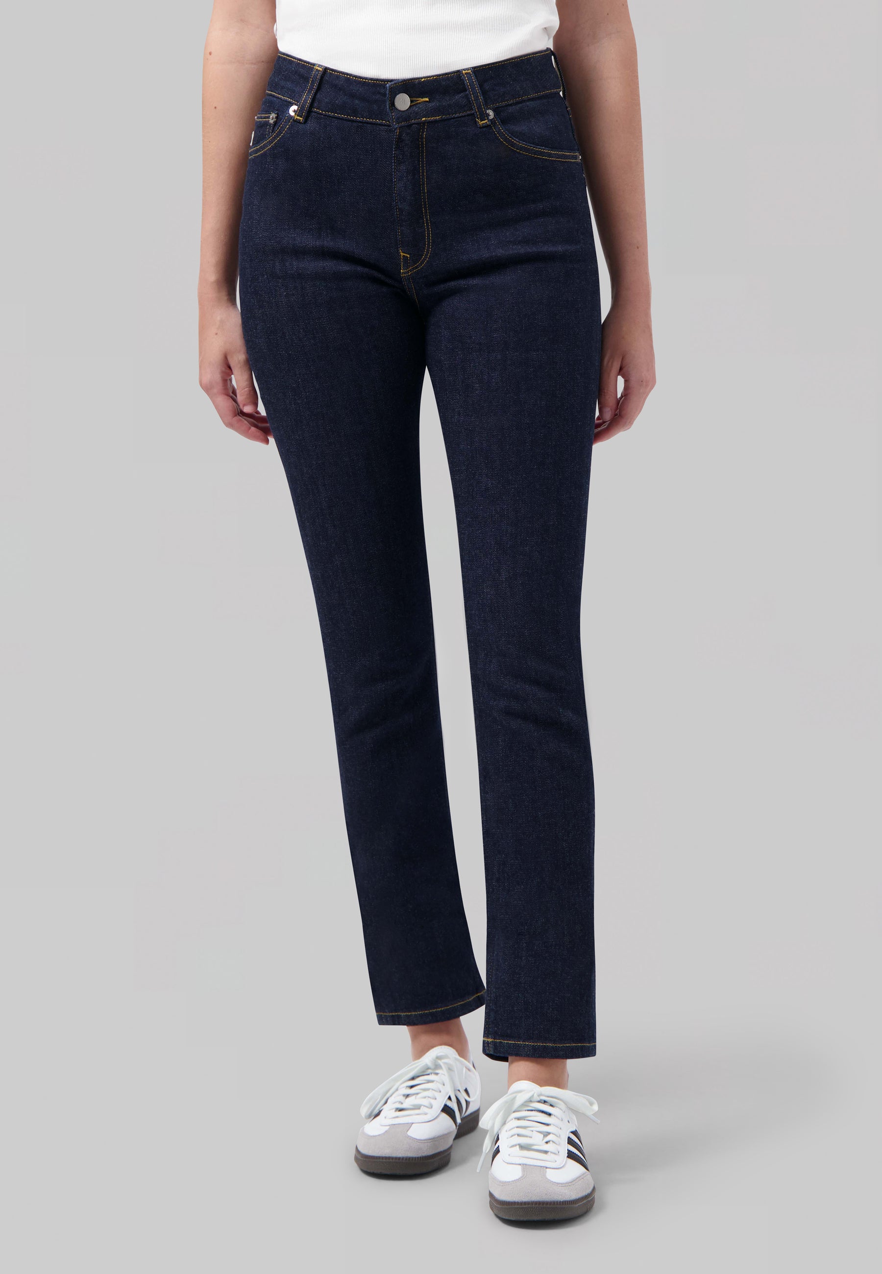 Dark blue high-waisted denim jeans with contrast stitching, worn with a white ribbed tank top and white sneakers.