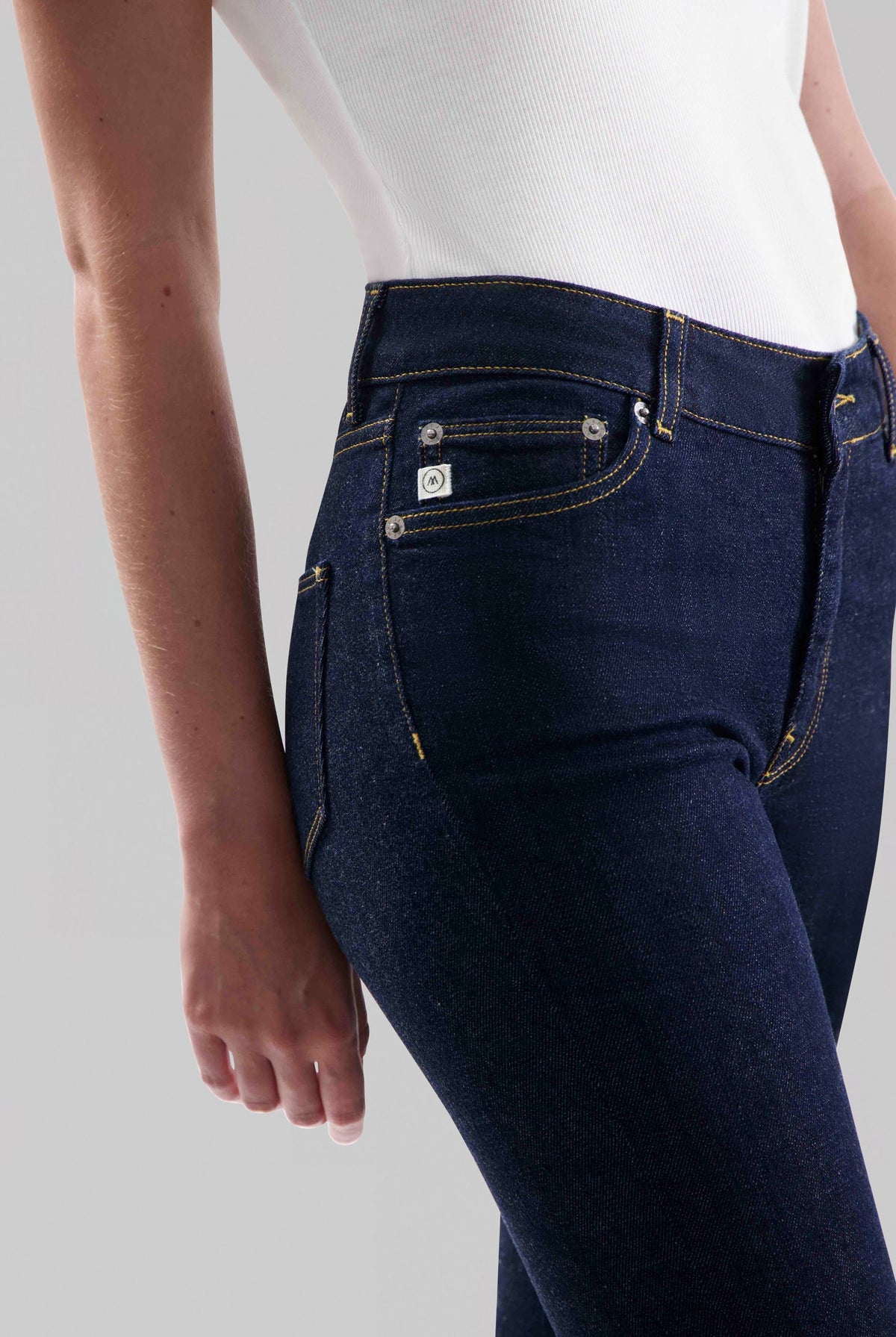 Close-up of dark indigo jeans with contrast stitching worn by model in a fitted white ribbed tank top, highlighting pocket details.
