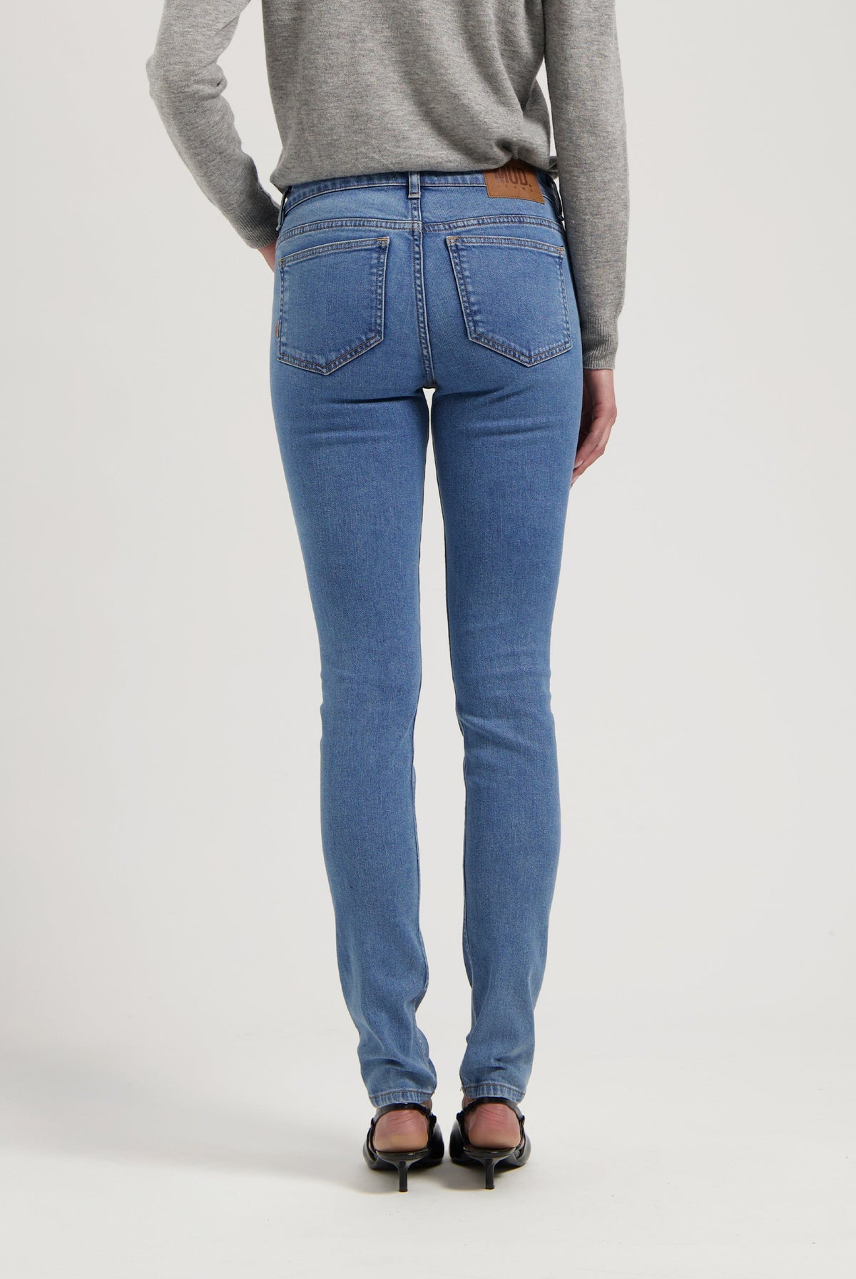 Slim-fit blue jeans with back pockets and a tan leather patch, paired with black heeled shoes and a gray sweater.