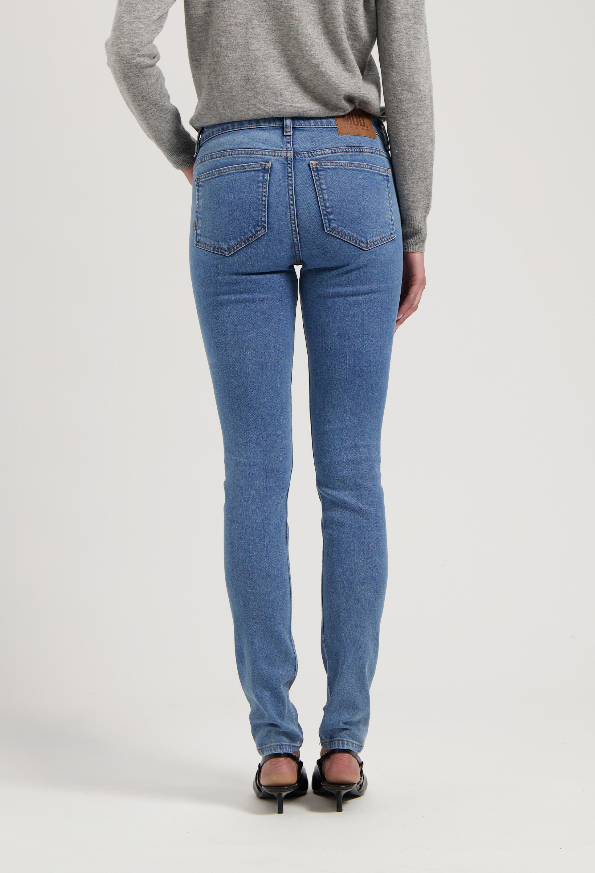 Slim-fit blue jeans with back pockets and a tan leather patch, paired with black heeled shoes and a gray sweater.