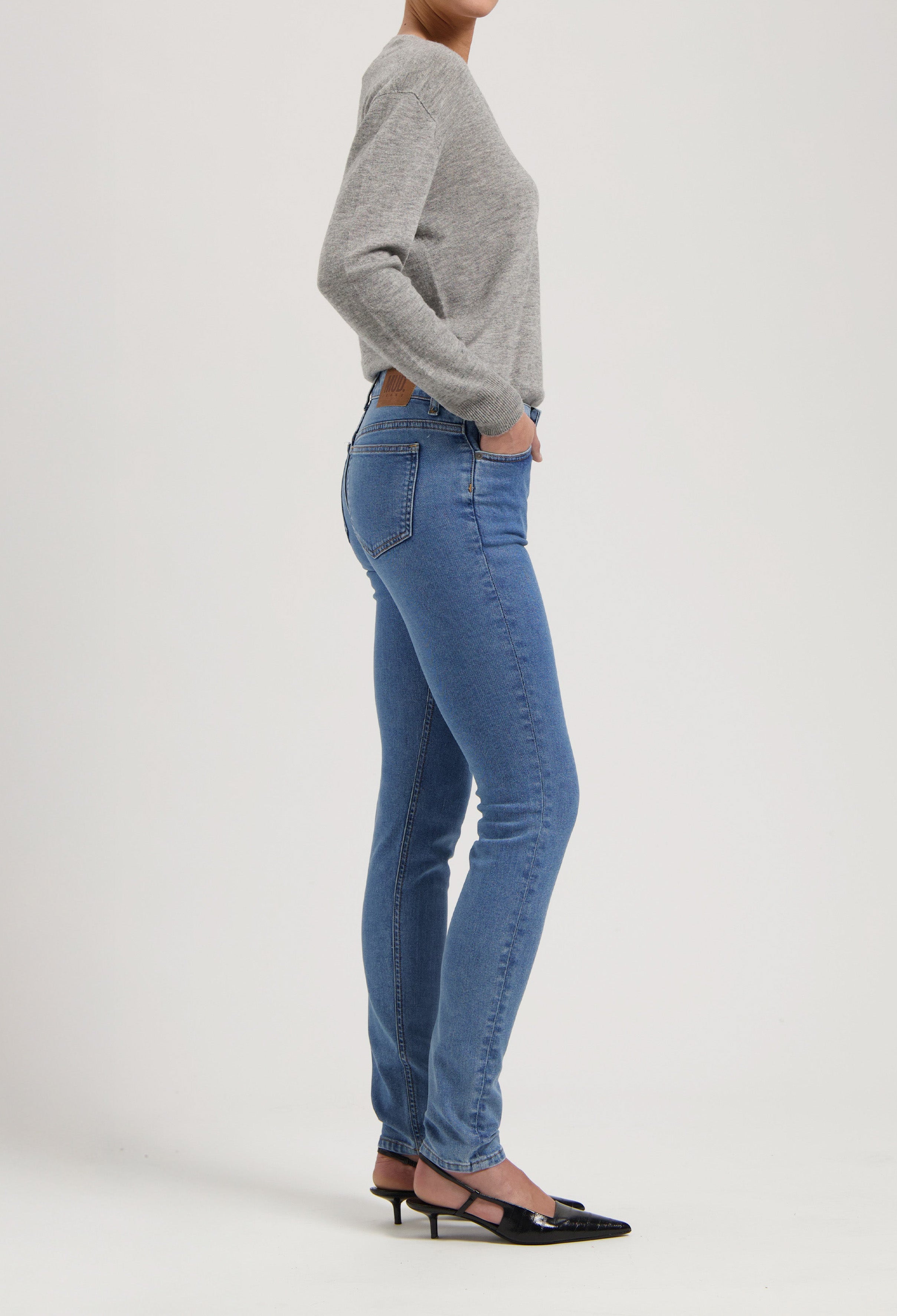 Woman wearing light blue denim jeans and gray sweater, side view, paired with black pointed-toe shoes against a white background.