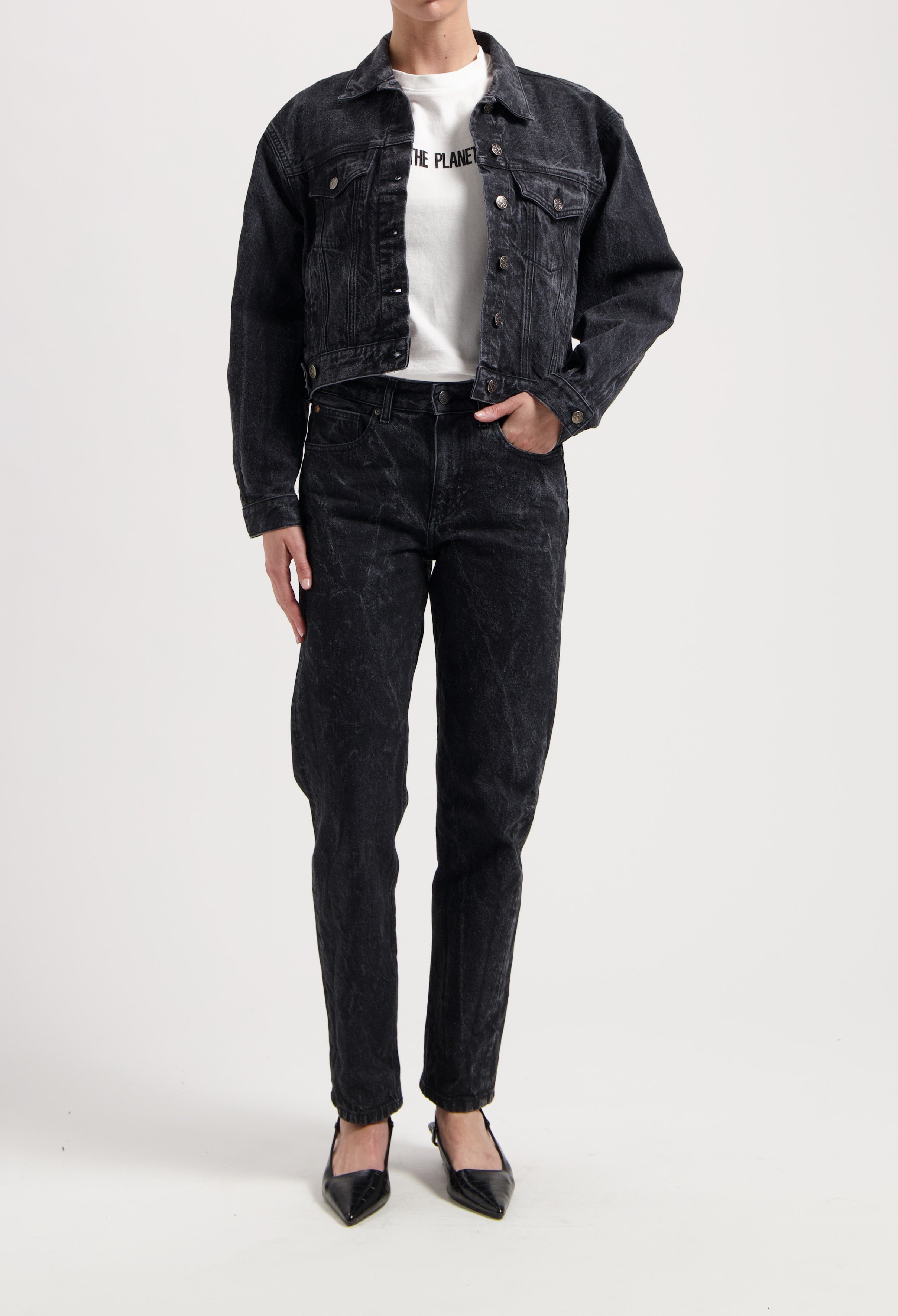 Model in black denim jacket and jeans, paired with a white graphic tee and pointed black heels, on a neutral background.