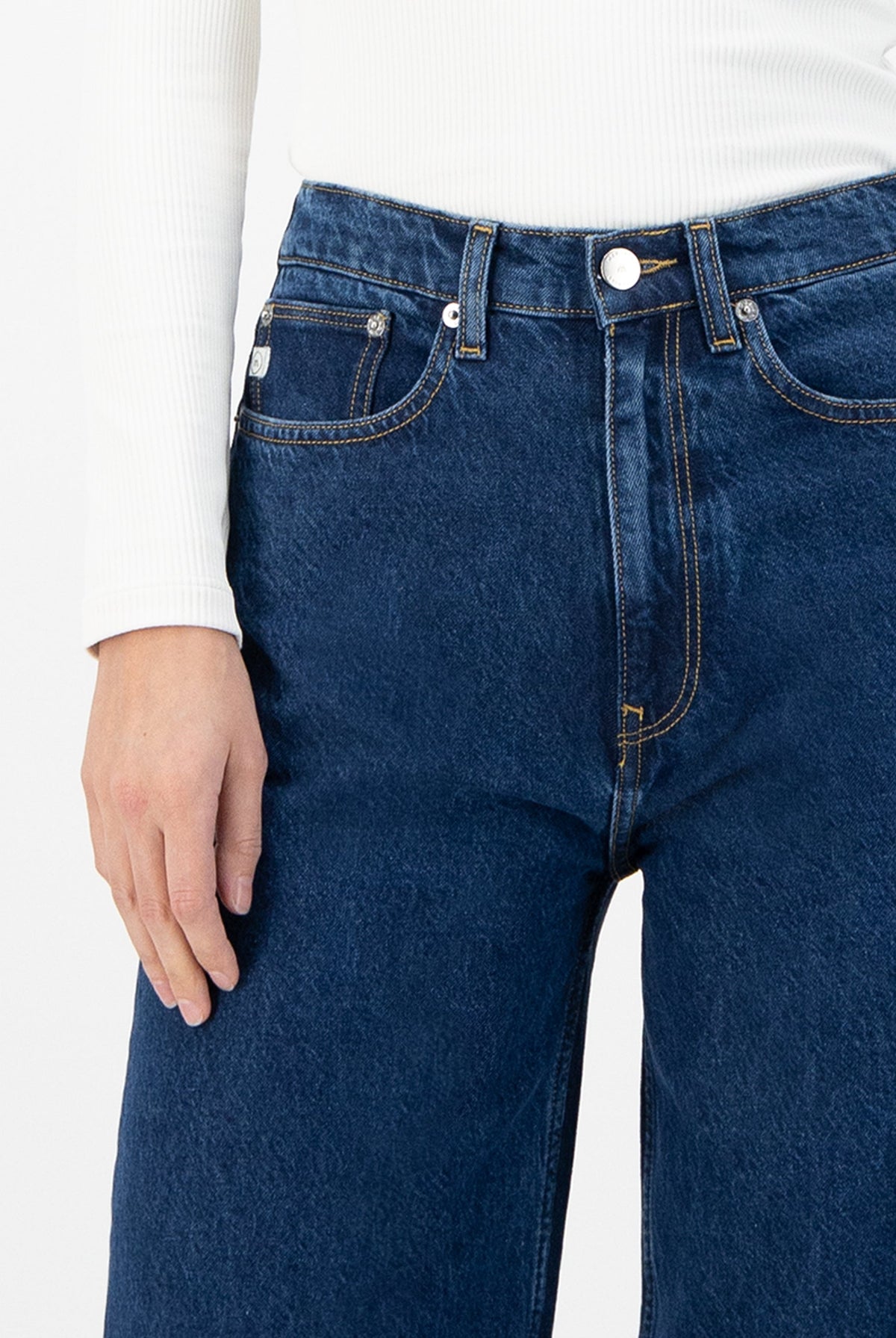 Close-up of blue high-waisted jeans with yellow stitching, worn with a white ribbed long-sleeve top.