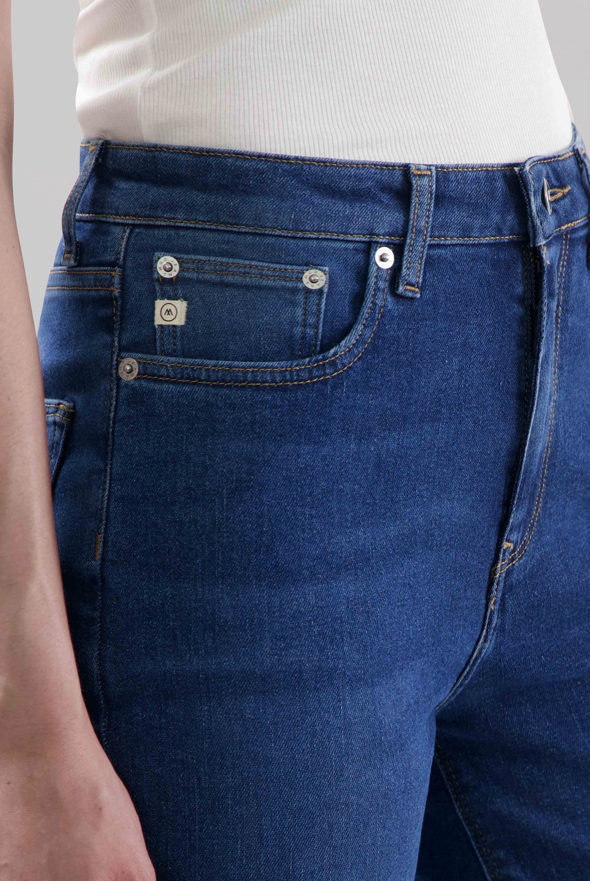 Dark blue denim jeans featuring front pockets and silver button details, worn with a white ribbed sleeveless top.