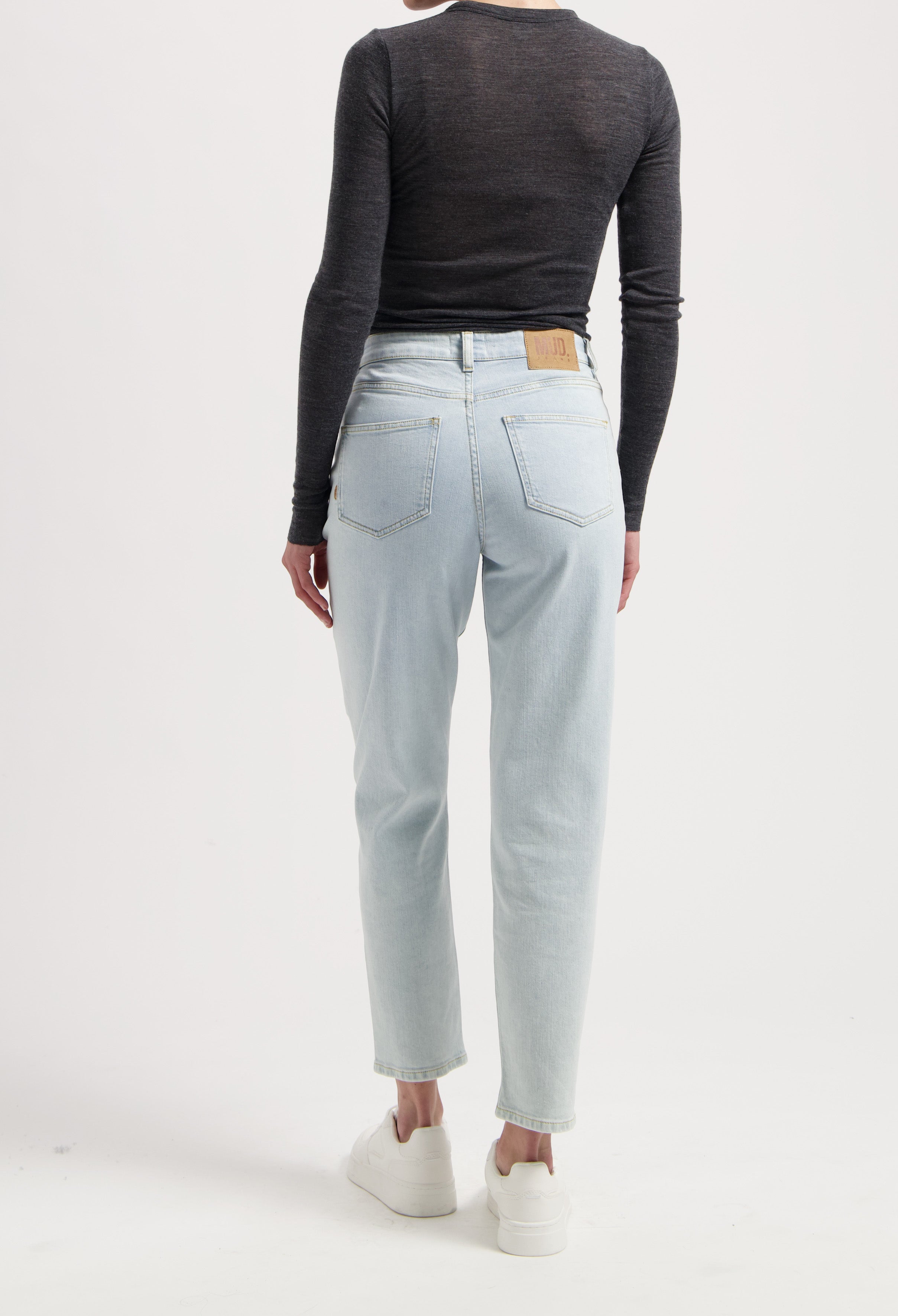 Light blue, high-waisted jeans from MUD Jeans, showcased with a black long-sleeve top and white sneakers in a studio setting.
