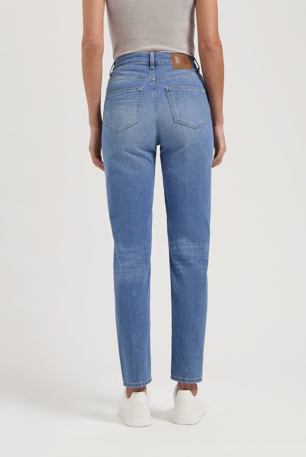 Mid-blue high-waisted jeans by MUD Jeans, featuring a tag on the waistband, paired with a gray sleeveless top and white sneakers.