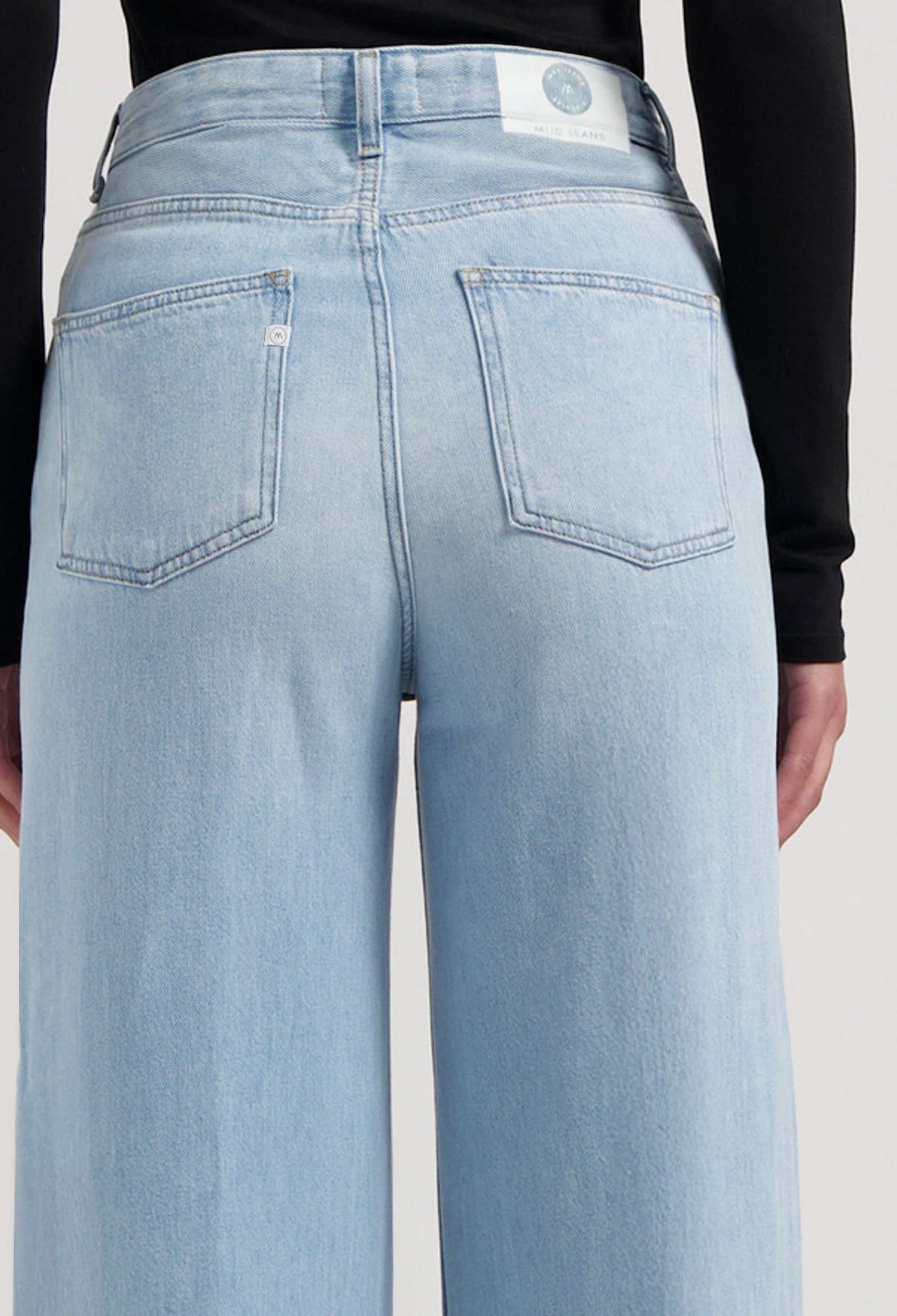 Rear view of light blue, high-waisted MUD Jeans in sustainable denim, showcasing a wide-leg fit paired with a sleek black top.