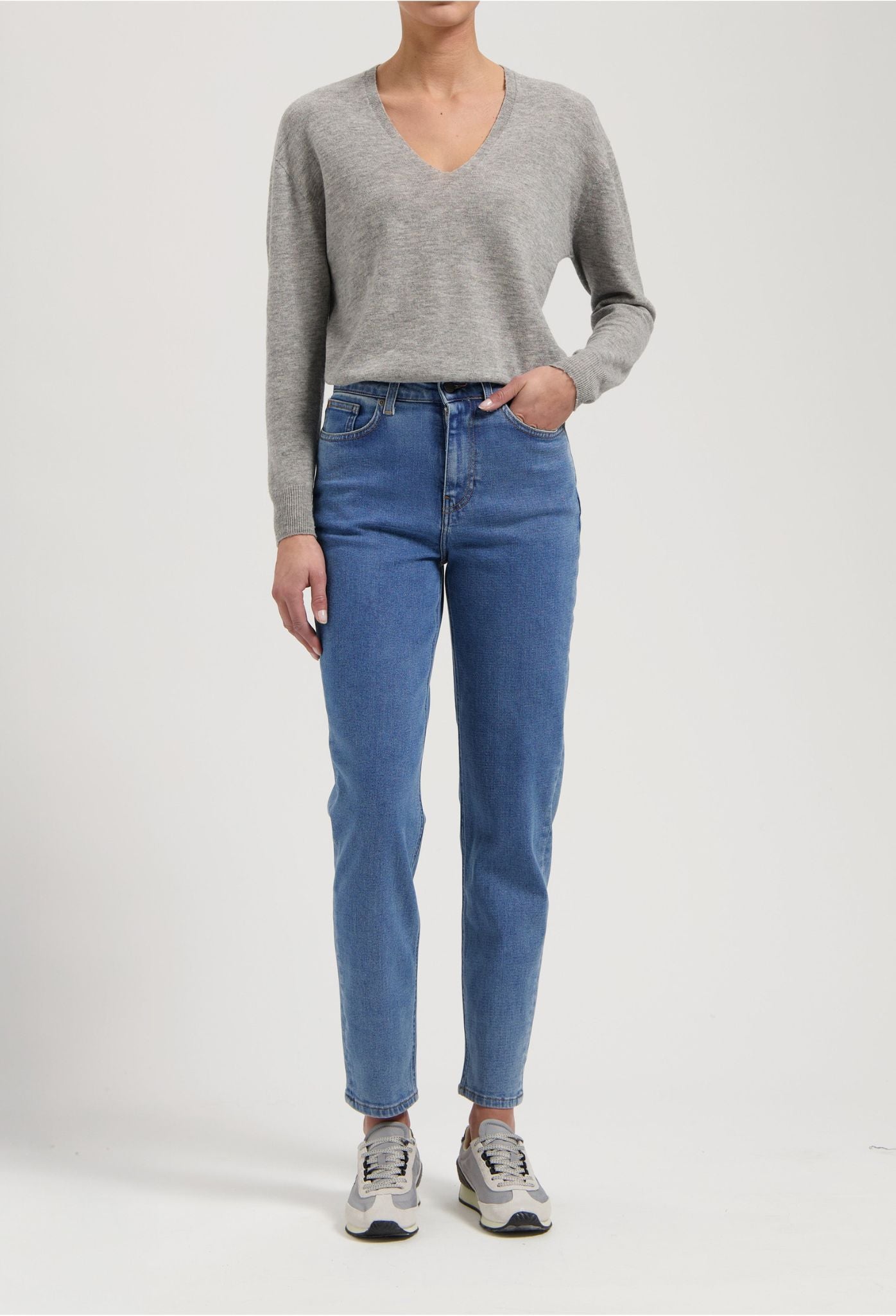 Woman wearing light blue straight-leg jeans and a gray V-neck sweater paired with casual gray sneakers.