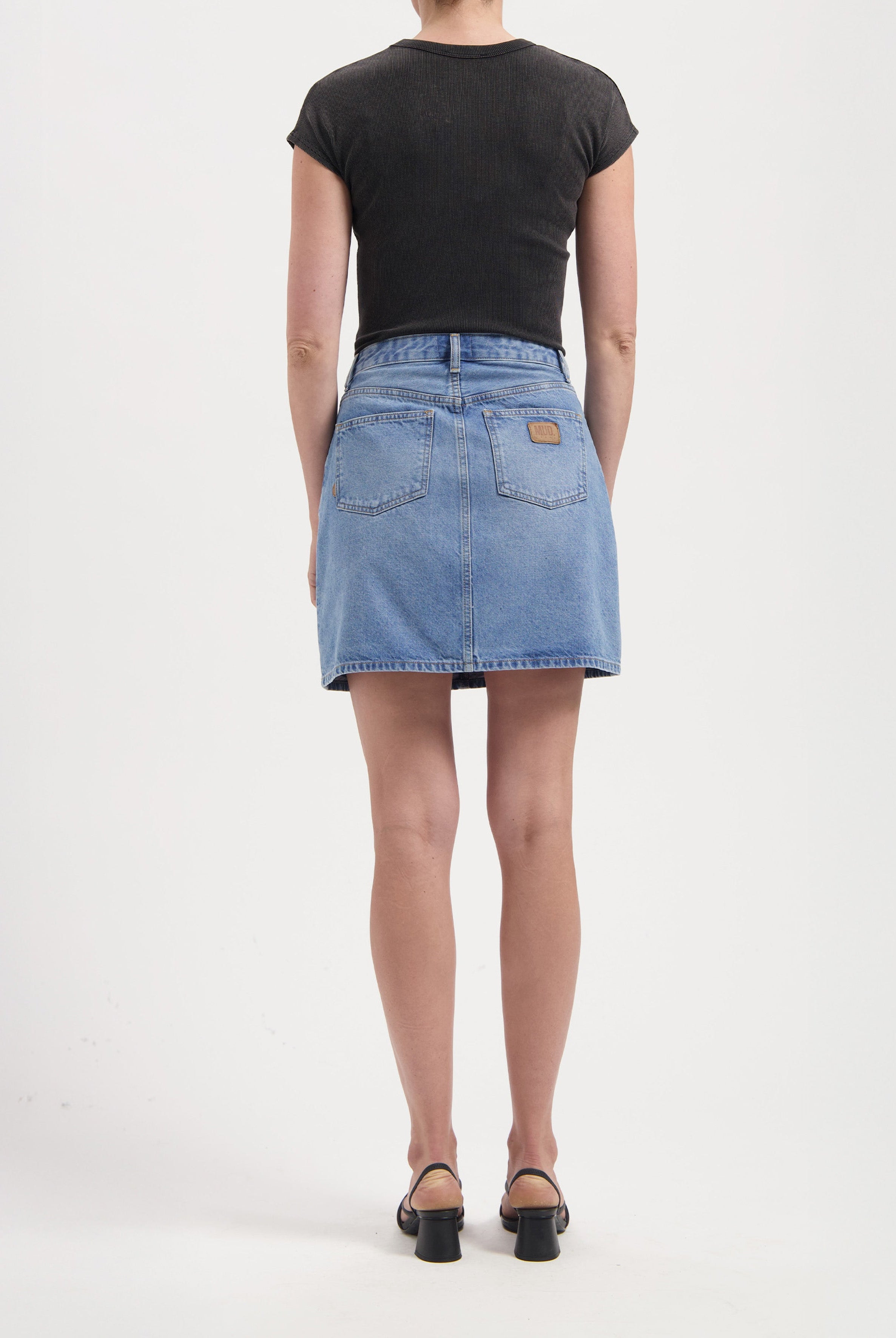 Back view of person wearing a blue denim skirt and black short-sleeve top paired with black heeled sandals.