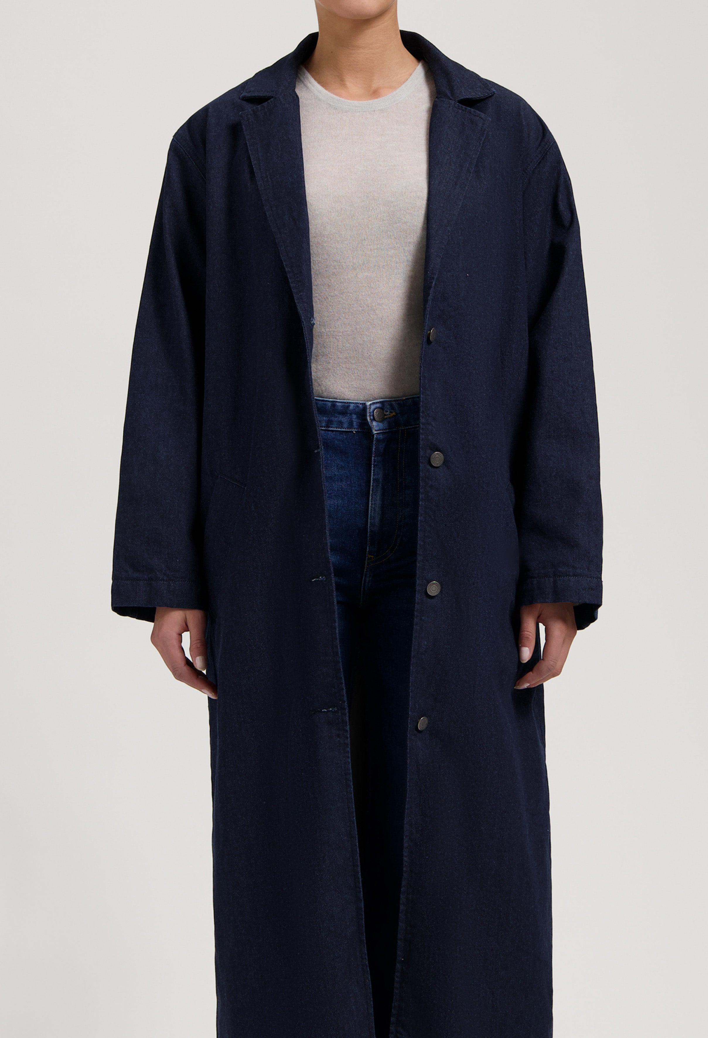 Long navy denim coat with button front, worn over a beige top and blue jeans, showcasing a minimalist casual style.