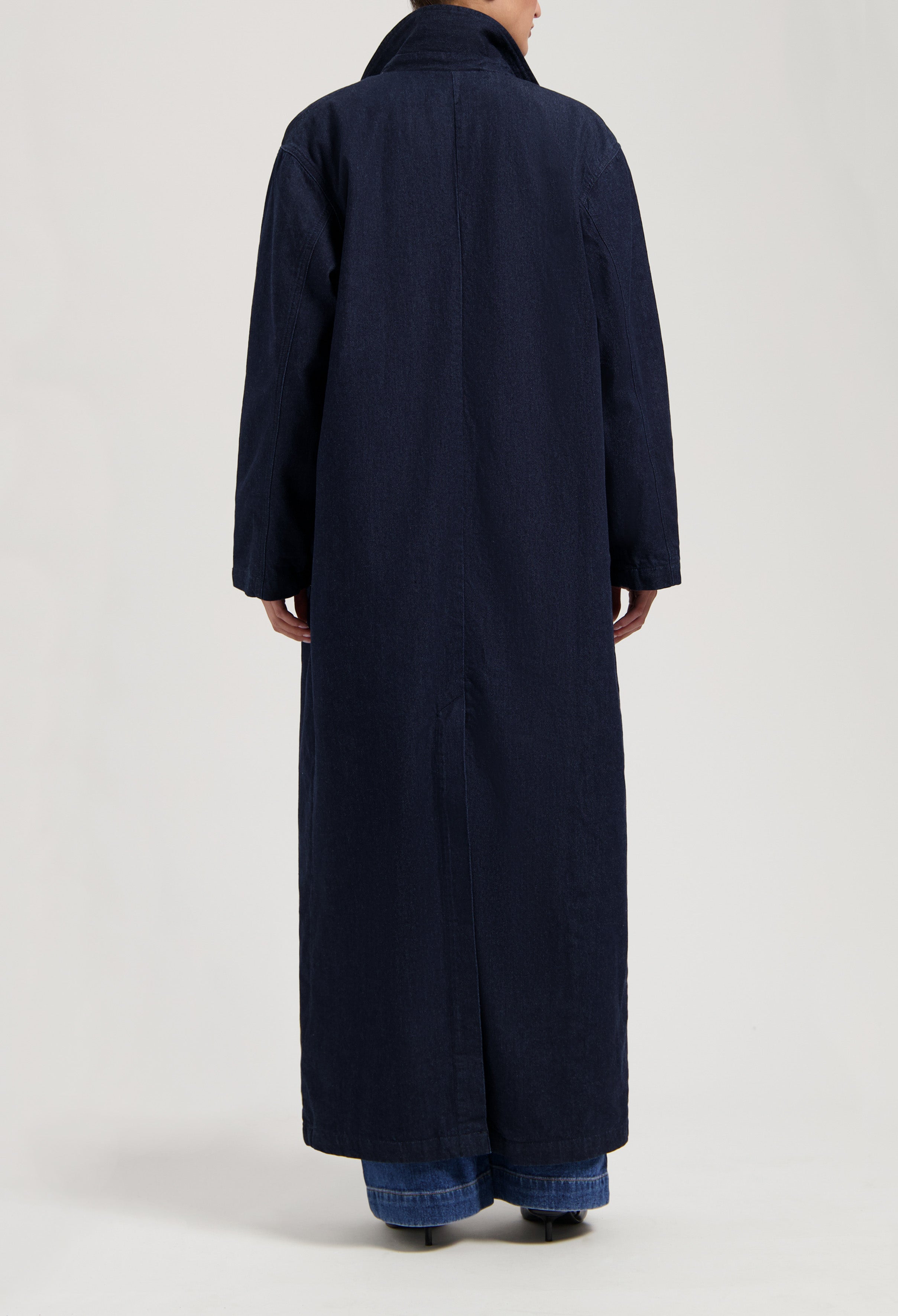 Dark blue, long denim overcoat with a high collar, shown from the back, worn over blue jeans for a sleek look.