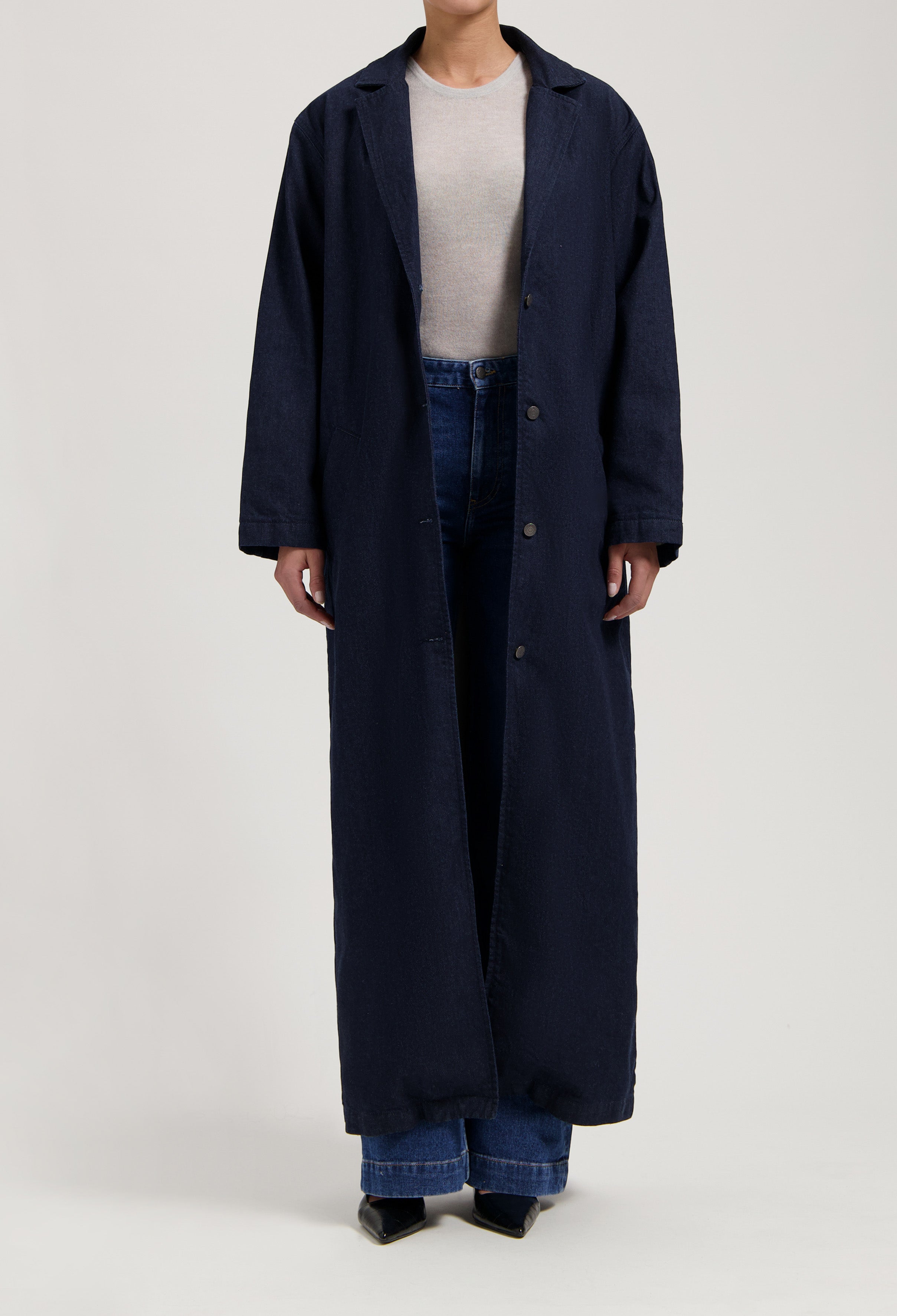 Long dark blue denim coat worn over light gray top and blue jeans, styled with black pointed-toe shoes.