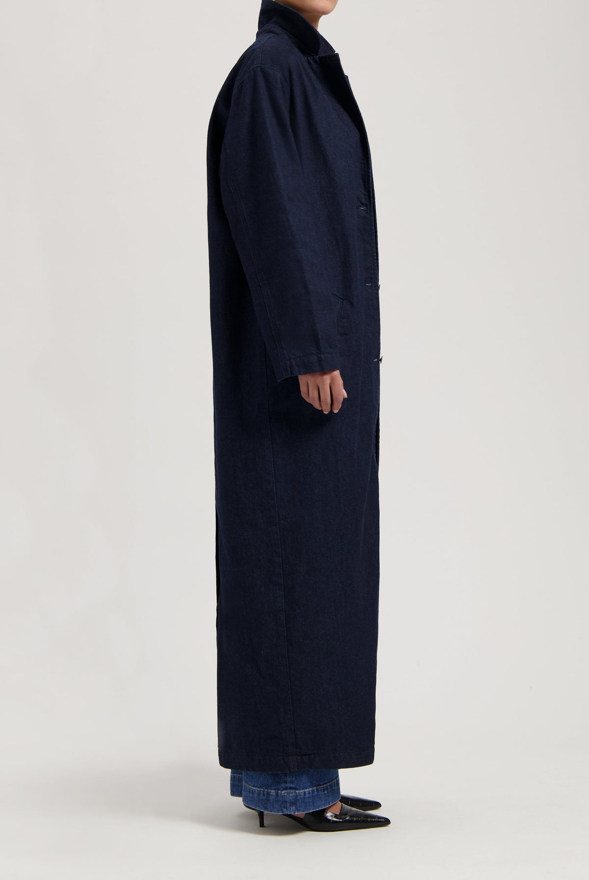 Long navy trench coat in dark fabric, styled with blue denim jeans and black pointed heels, side view.