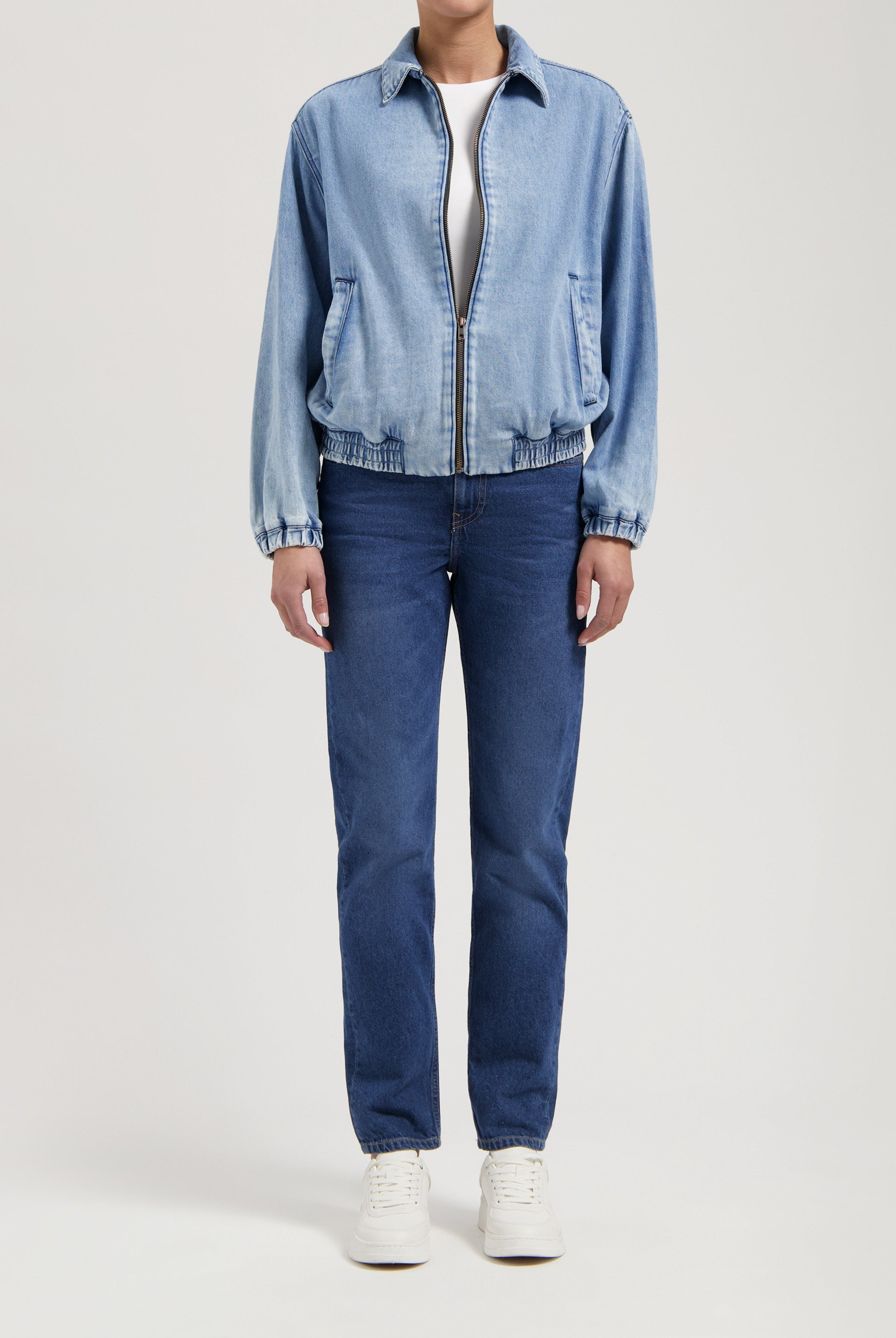 Person wearing a light blue denim jacket with front zipper, paired with dark blue straight-leg jeans and white sneakers.
