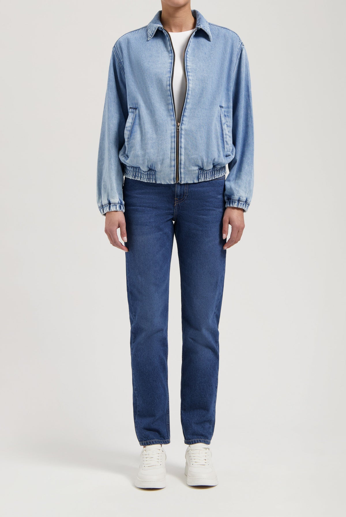 Person wearing a light blue denim jacket with front zipper, paired with dark blue straight-leg jeans and white sneakers.