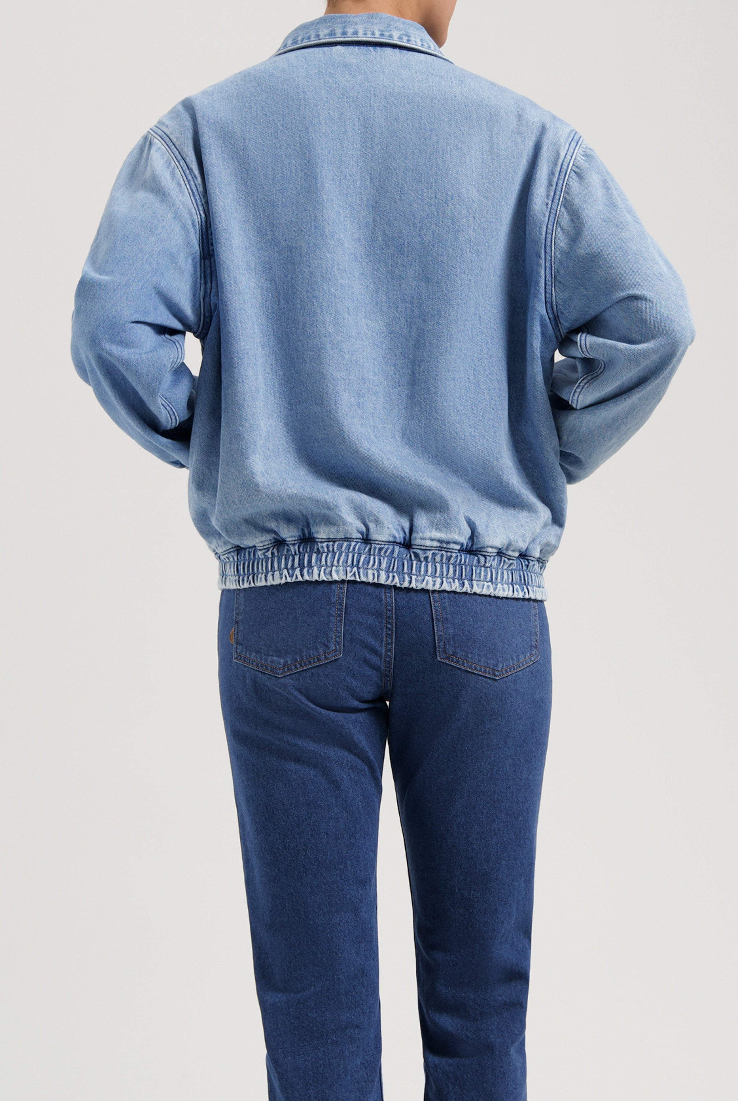 Light blue denim jacket with elasticated hem and long sleeves, paired with dark blue straight-leg jeans, showing back view.