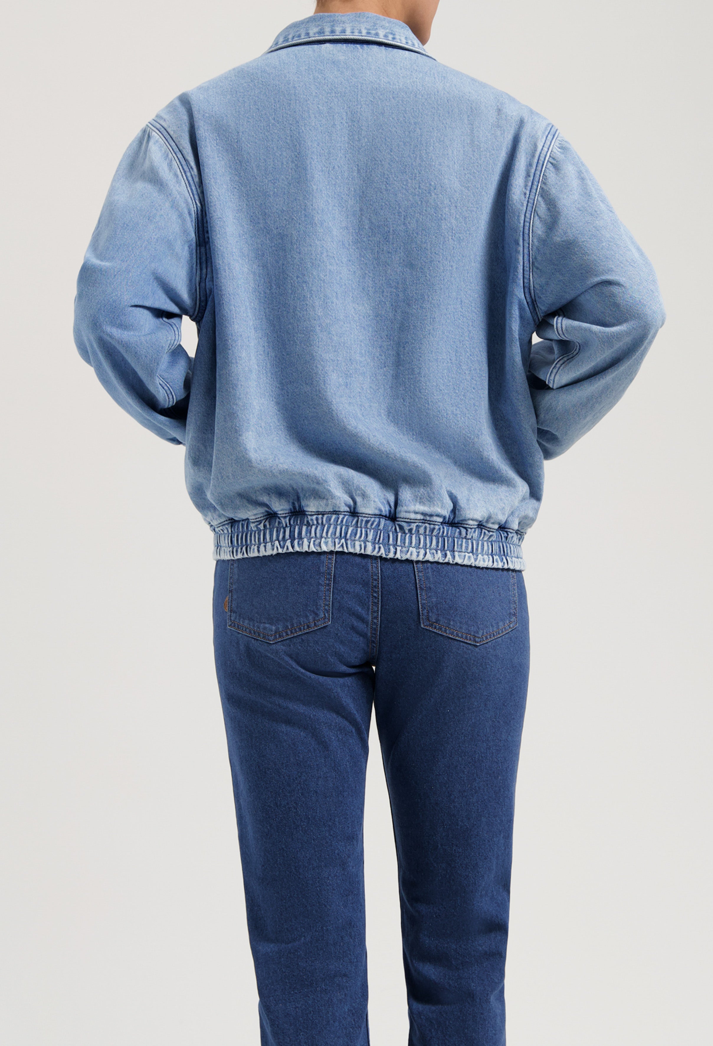 Light blue denim jacket with elasticated hem and long sleeves, paired with dark blue straight-leg jeans, showing back view.