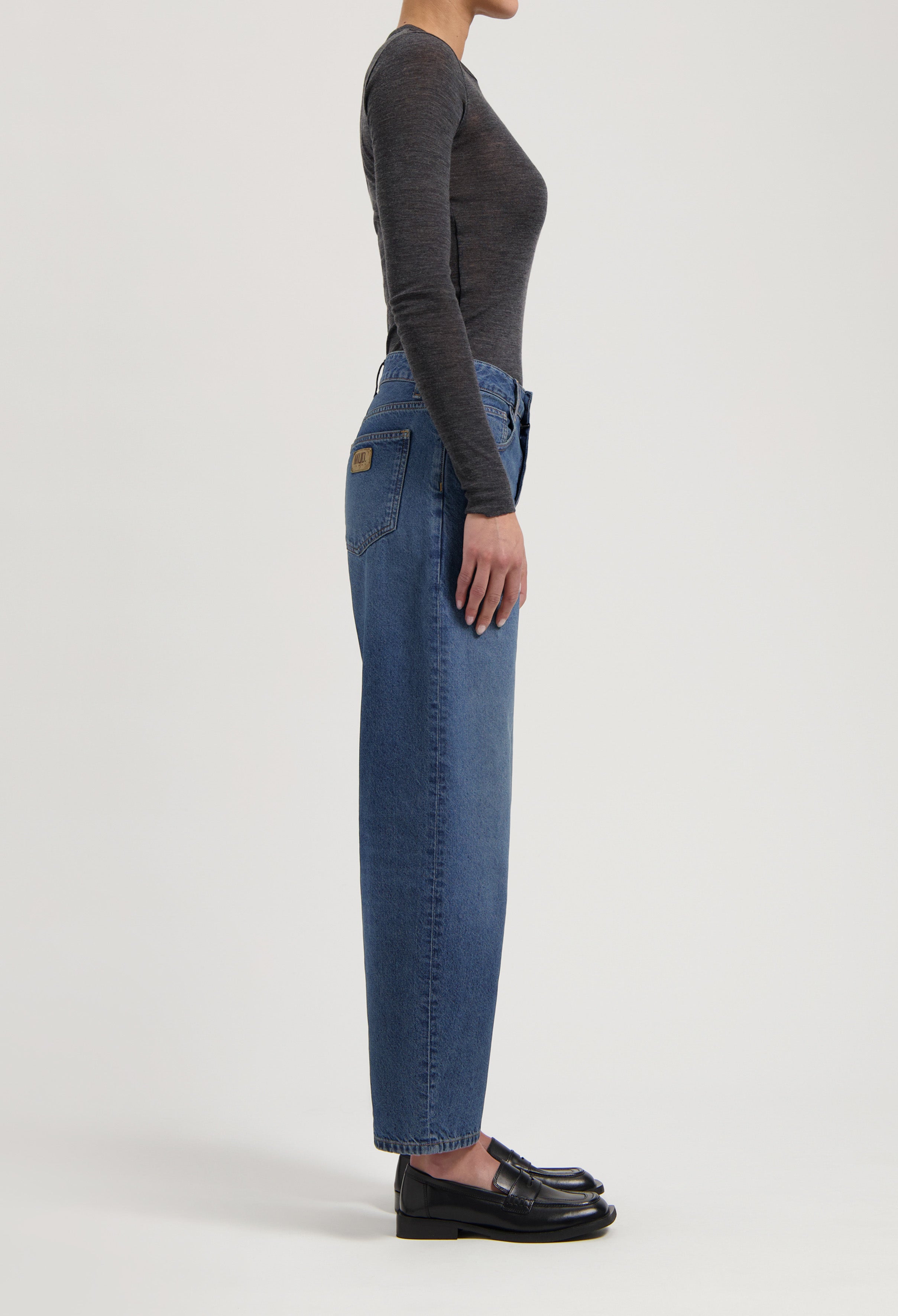 Side view of a person wearing high-waisted blue jeans and a fitted long-sleeve gray top paired with black leather loafers.