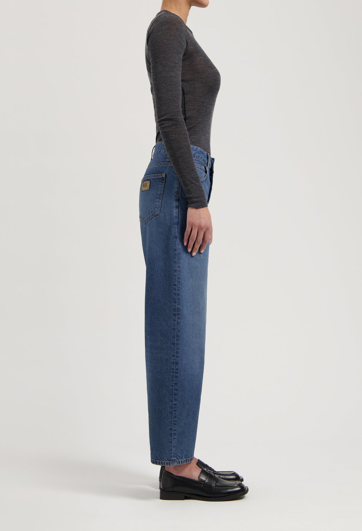 Side view of a person wearing high-waisted blue jeans and a fitted long-sleeve gray top paired with black leather loafers.