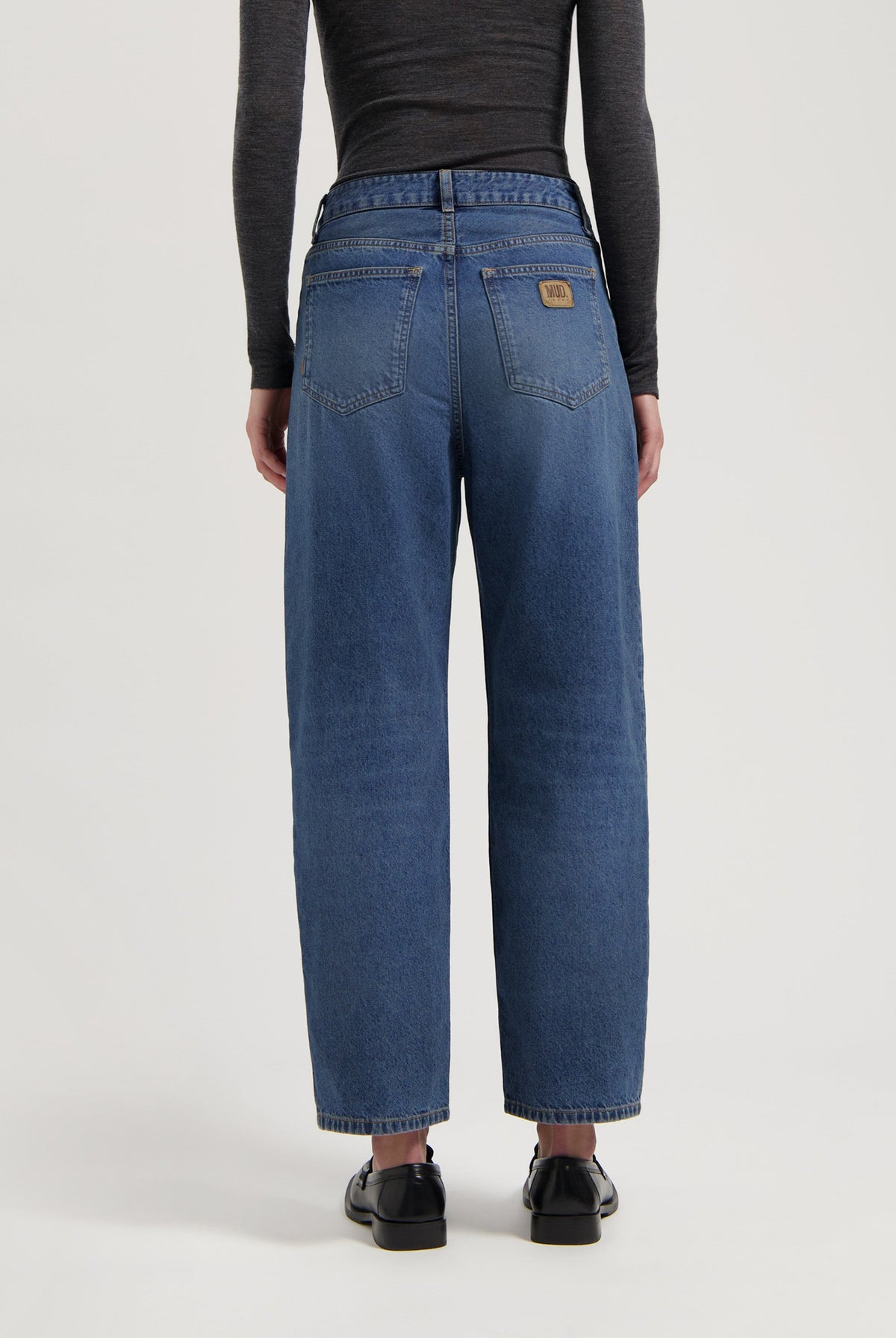 Loose-fit, mid-blue jeans by MUD Jeans made from organic cotton, styled with a dark long-sleeve top and black shoes.
