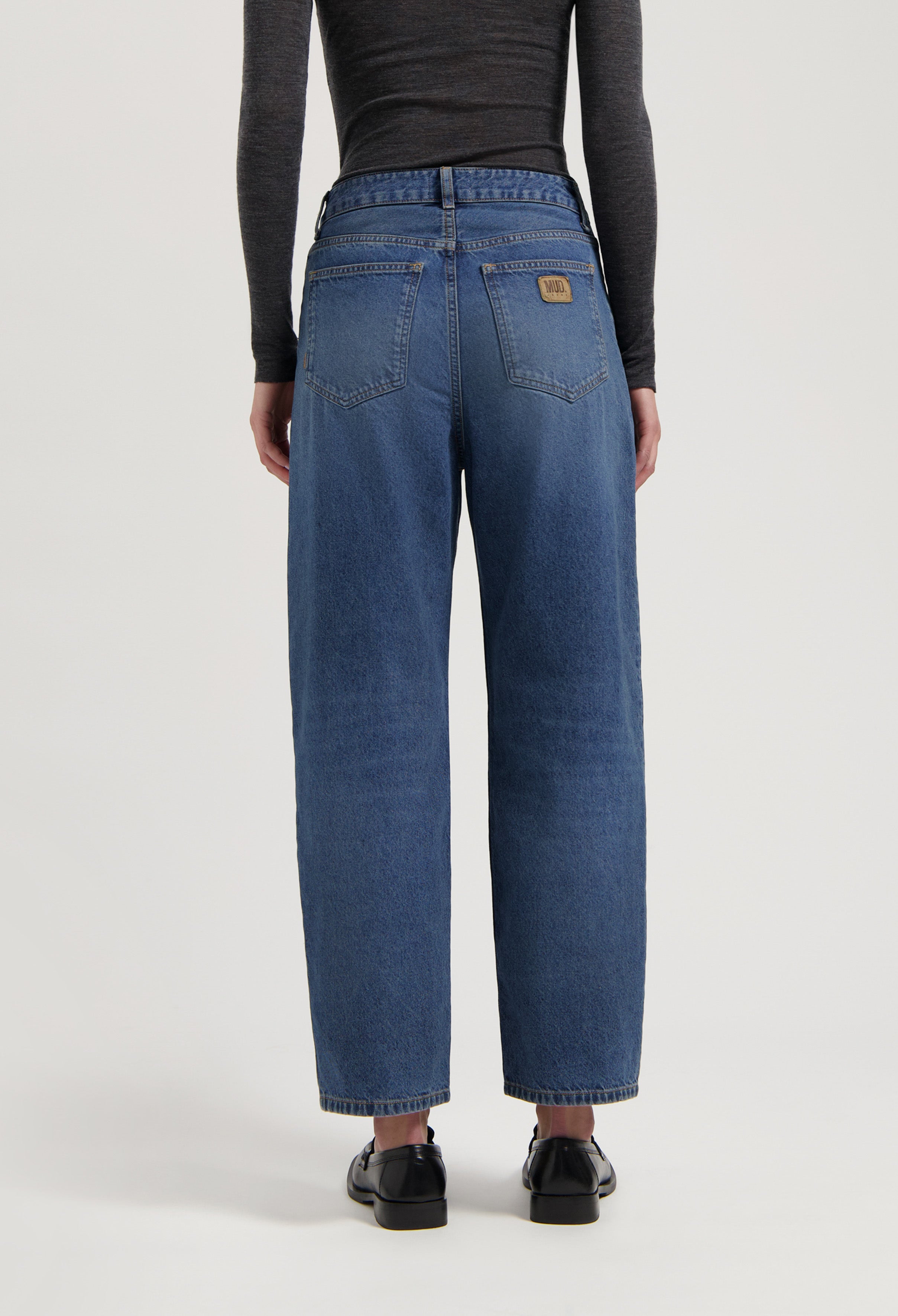 Loose-fit, mid-blue jeans by MUD Jeans made from organic cotton, styled with a dark long-sleeve top and black shoes.